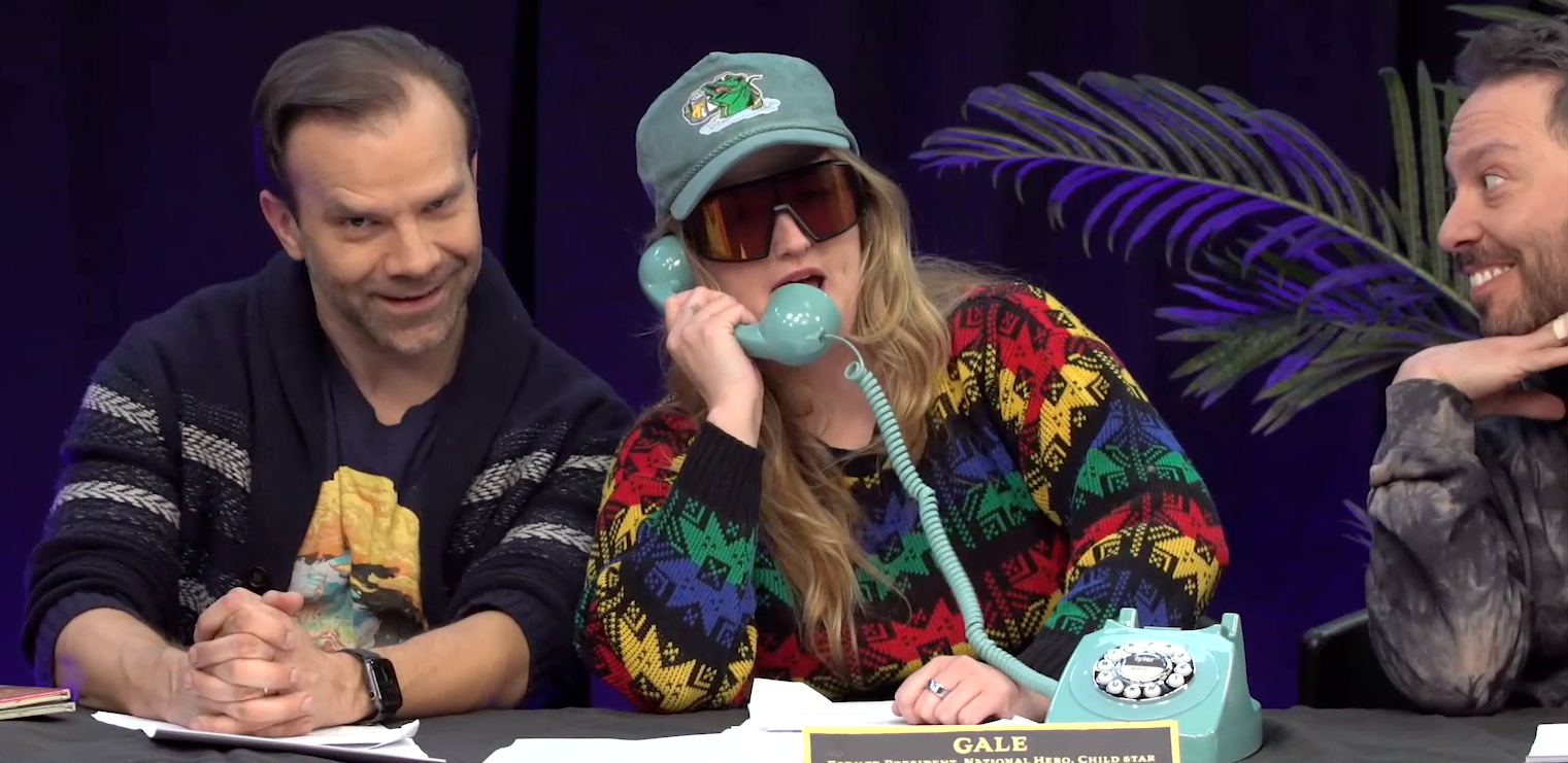 The Critical Role Announcement Playhouse table. Ashley, dressed as Gale, speaks into a sky blue rotary phone. Liam sits to her right, head cocked and face skewed into a mischievous smile. Ashley wears visor sunglasses and a blue baseball cap with an image of a large green fish drinking a stein of beer. She has on a sweater in blacks and primary colors. On the table in front of the phone is a name plate reading “GALE: FORMER PRESIDENT, NATIONAL HERO, CHILD STAR”. Sam looks on, smiling gleefully from the right edge of the image. 