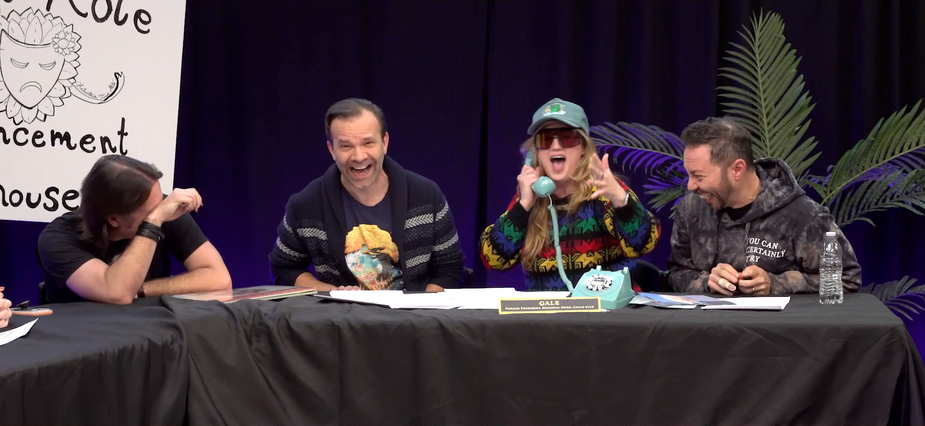 The Critical Role Announcement Playhouse table. Ashley, dressed as Gale, speaks emphatically into a sky blue rotary phone while Matt, Liam, and Sam erupt in uncontrolled laughter. Ashley wears visor sunglasses and a blue baseball cap with an image of a large green fish drinking a stein of beer. She has on a sweater in blacks and primary colors. On the table in front of the phone is a name plate reading “GALE: FORMER PRESIDENT, NATIONAL HERO, CHILD STAR”.