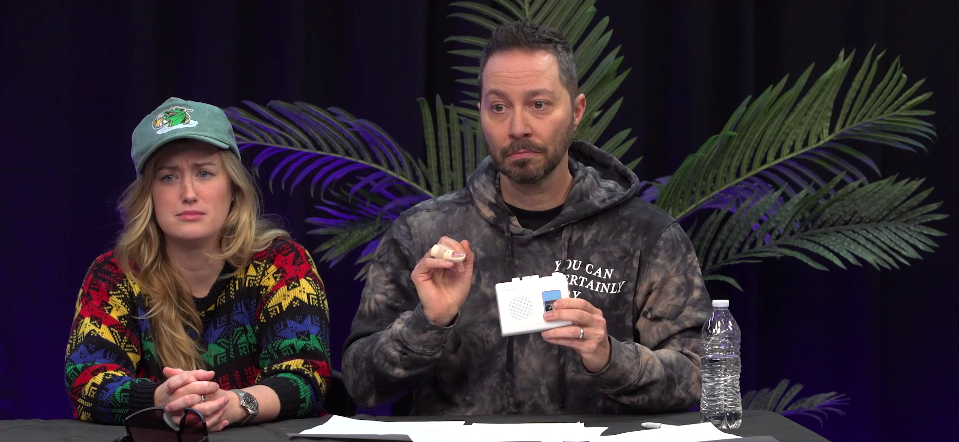 The Critical Role Announcement Playhouse table. Sam holds a small white cassette player with the play button pressed down. His face and body display discomfort as he listens. Ashley sits next to him looking confused, her hands clasped on the table in front of her. Ashley wears a blue baseball cap with an image of a large green fish drinking a stein of beer and a sweater in blacks and primary colors. Sam wears a gray hoodie with the text “YOU CAN CERTAINLY TRY” in white over the left breast. A large potted plant is visible in the background and a plastic disposable water bottle sits in front of Sam.