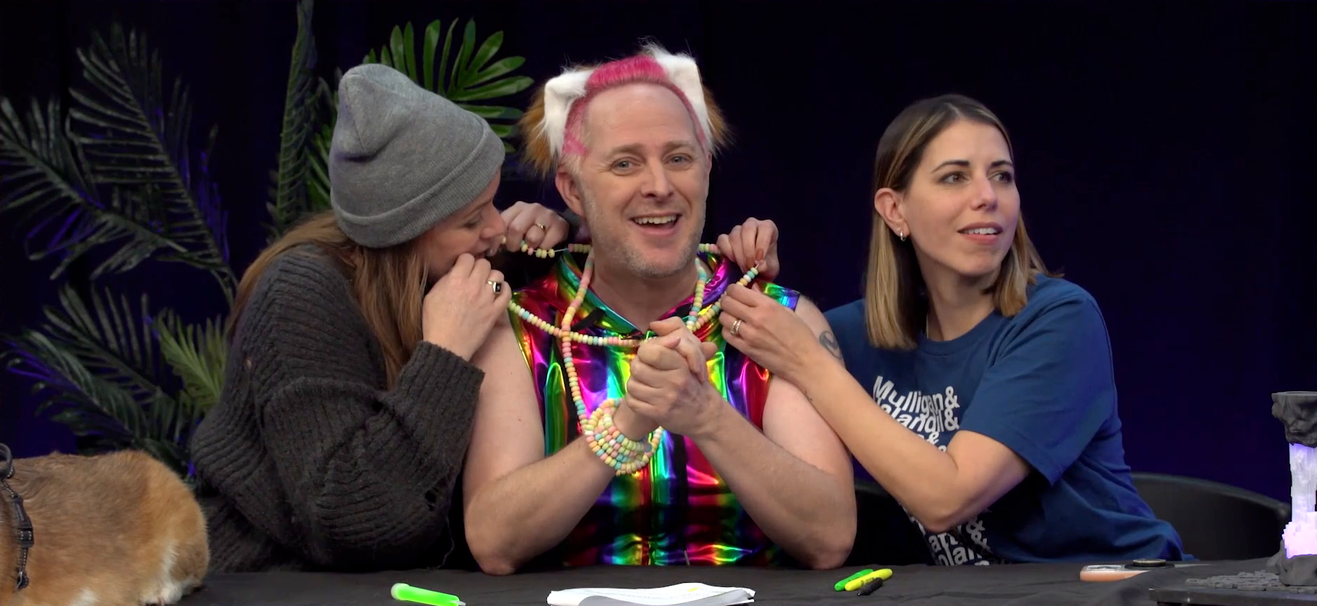 The Critical Role Announcement Playhouse table. Taliesin is dressed in a bright rave outfit and candy necklaces, while Marisha and Laura on either side of him eat from those necklaces. Taliesin wears a metallic rainbow tanktop and a pink, white, and brown wig, and clasps his hands in front of him in good-natured discomfort. Marisha grabs one end of a necklace in both hands and chomps down on it, while Laura holds the other end of the necklace, looking off screen, smiling and chewing.