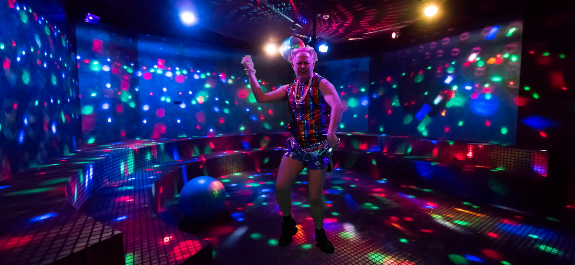Taliesin dancing uncomfortably, green-screened onto an empty rave dancefloor. He wears a metallic rainbow tanktop, short metallic shorts, and a number of candy necklaces, and carries a green glow stick in each hand. The green-screened background has a disco ball, a scattering of colorful lights across the walls and floor, a blue sphere sitting on the ground, and tiled bench-like platforms lining the edges of the room.