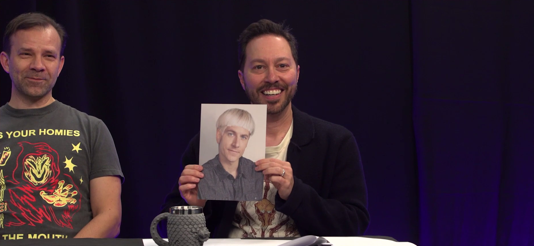 The Critical Role Announcement Playhouse table. Sam holds up a photograph of Matt with a platinum blond bowlcut. Matt in the image looks forward with a neutral expression. Sam smiles while holding the photograph. Liam sits next to him, holding in a laugh. Liam wears a shirt with an image of a grim reaper reading “KISS YOUR HOMIES ON THE MOUTH”. A mug with a gray dragonscale texture sits in front of Sam.