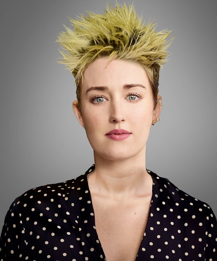 A headshot of Ashley with spiky hair dyed dandelion yellow and brown roots, in a punk style. The hair is short-cut. Ashley looks directly at the viewer placidly. She wears a V-neck black blouse with white polkadots. She has pink lipstick, two gold ring piercings towards the top of her left ear and one gold piercing on the lobe. The picture is set against a solid gray background.