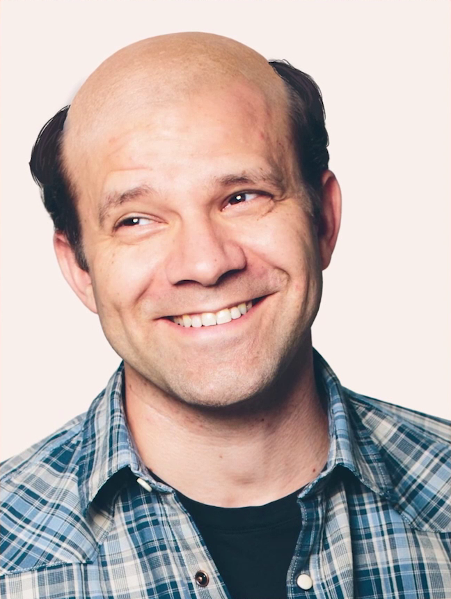 A headshot of Liam fully bald except for short brown hair on the sides of his head. He smiles widely and genially, head tilted, looking up and to the right. He wears a blue and white plaid collared button-up shirt with fastening snaps over a black undershirt. The picture is set against a solid white background.