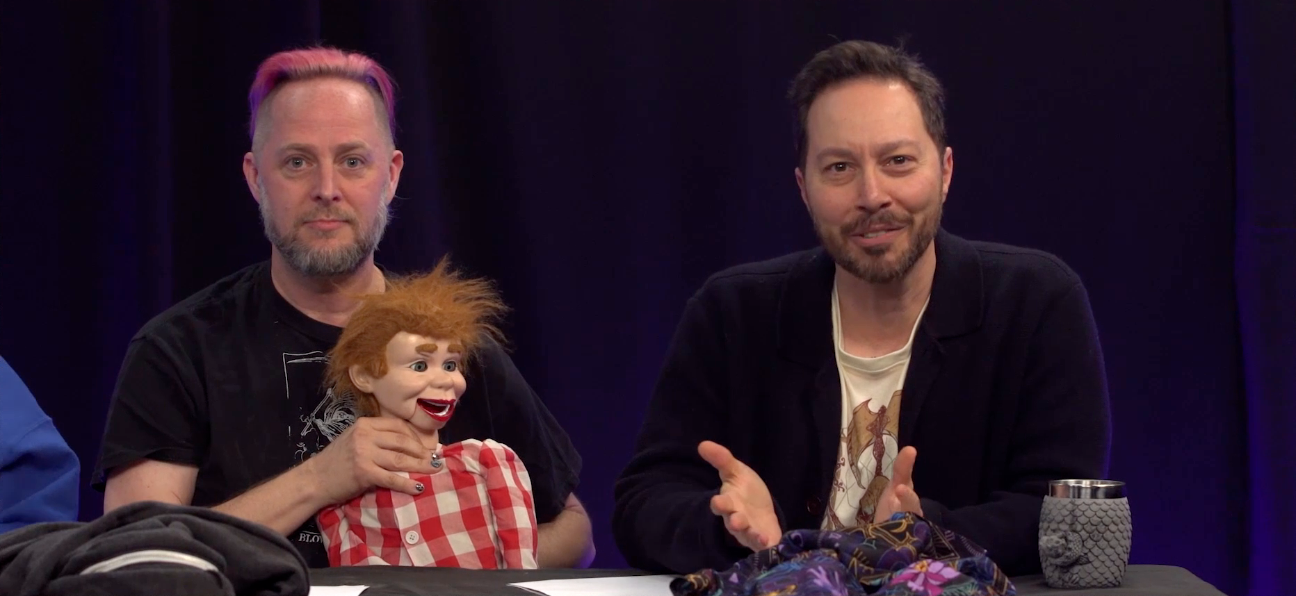 Taliesin and Sam sitting at the Critical Role Announcement Playhouse table. Taliesin holds a ventriloquist dummy with a plaid red and white shirt and messy ginger hair. One hand is behind the dummy’s back and the other rests on its neck. The dummy’s mouth is open as if talking. Taliesin and Sam both stare directly at the camera. Sam gestures forward, as if explaining something, while his face looks confused. Taliesin’s face is creepily passive.