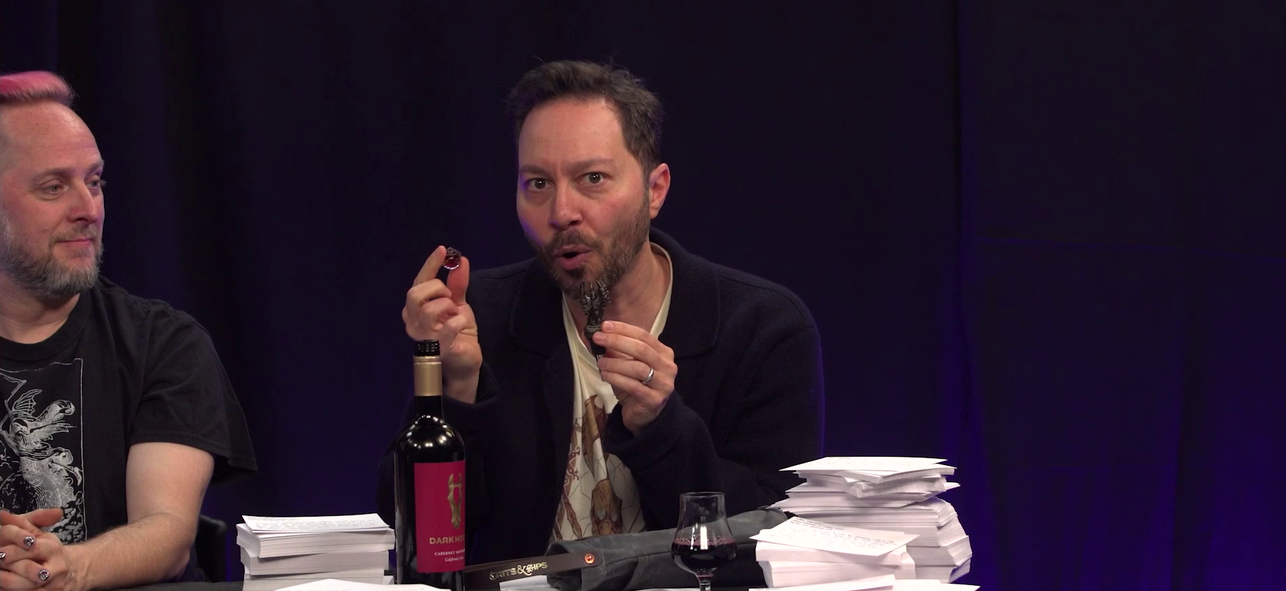 Sam and Taliesin sitting at the Critical Role Announcement Playhouse table. Sam holds a red translucent 20-sided die in his right hand and a claw-shaped wine stopper in his left hand. His mouth is in an O shape as if giving an exaggerated pitch. On the table in front of him is an open wine bottle with the text “DARK HORSE” and a gold image of a horse head on the label. Next to the bottle is a glass partially filled with red wine, a wine bottle tote reading “CRITS & SIPS” on the handle, and several large stacks of hundreds of index cards. Taliesin looks over at Sam, vaguely amused.