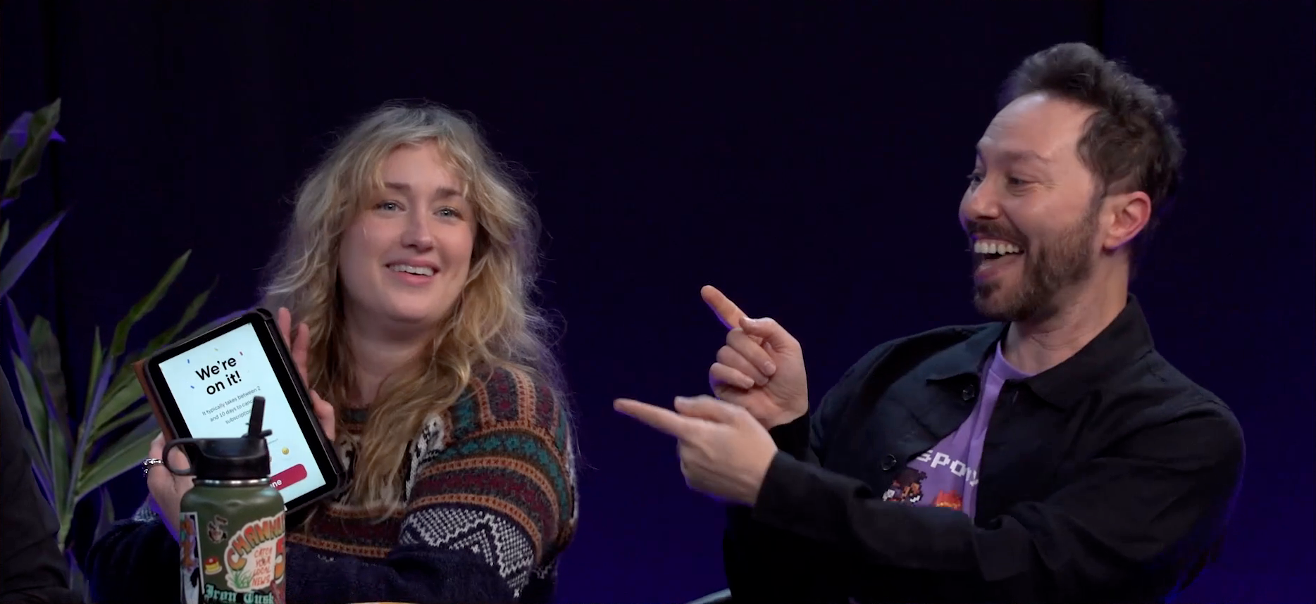 Ashley and Sam sitting at the Critical Role Announcement Playhouse table. Ashley holds up a tablet with the text “We’re on it!” surrounded by confetti. Below reads “It typically takes between 2 and 10 days to cancel a subscription”, and a red “Done” button is at the bottom of the screen beneath multiple happy face emojis. Ashley smiles as she displays the tablet, and Sam gives an open-mouth grin, hands raised to point at Ashley.