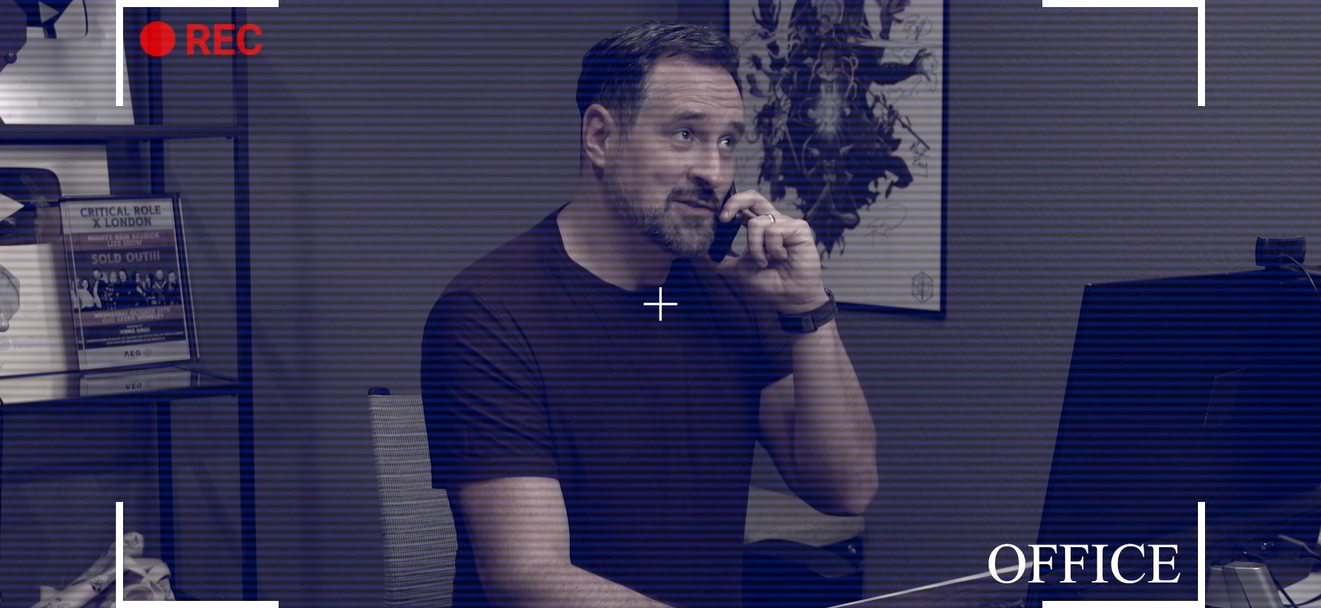 A CCTV-style video frame showing Travis sitting at his desk talking on his phone and smiling tenderly. A red “recording” icon is at the top left, the bottom right of the frame is labeled “OFFICE”, and horizontal lines run across the frame similar to a CRT monitor. On the desk is a computer monitor with a webcam mounted on top. On the wall behind Travis is a Mighty Nein poster with all the cast’s signatures on it. On a shelf behind Travis is a YouTube views award and a plaque for the Mighty Nein Reunion Live Show in London, with “SOLD OUT!!!” in large text over an image of the cast.