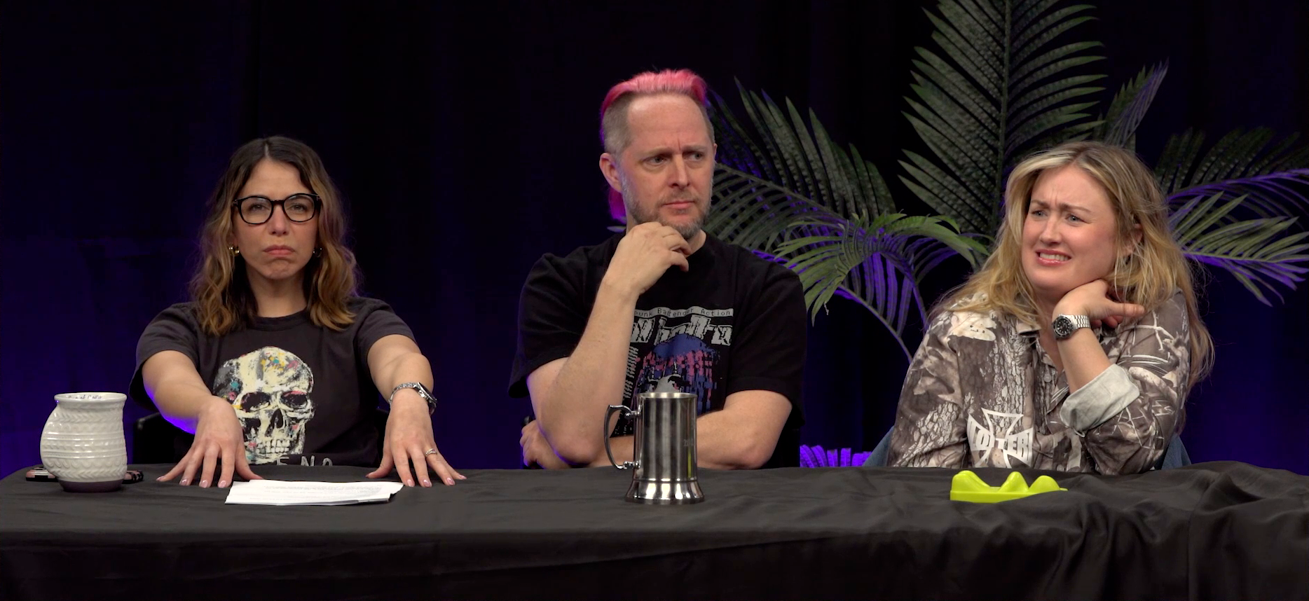 Laura, Taliesin, and Ashley sitting at the Critical Role Announcement Playhouse table. Laura looks upset, her fingers pressing on the table in front of her and her face tense and angry. Taliesin’s hand is on his chin, looking thoughtful and confused. Ashley grimaces, looking very worried about something she’s hearing.