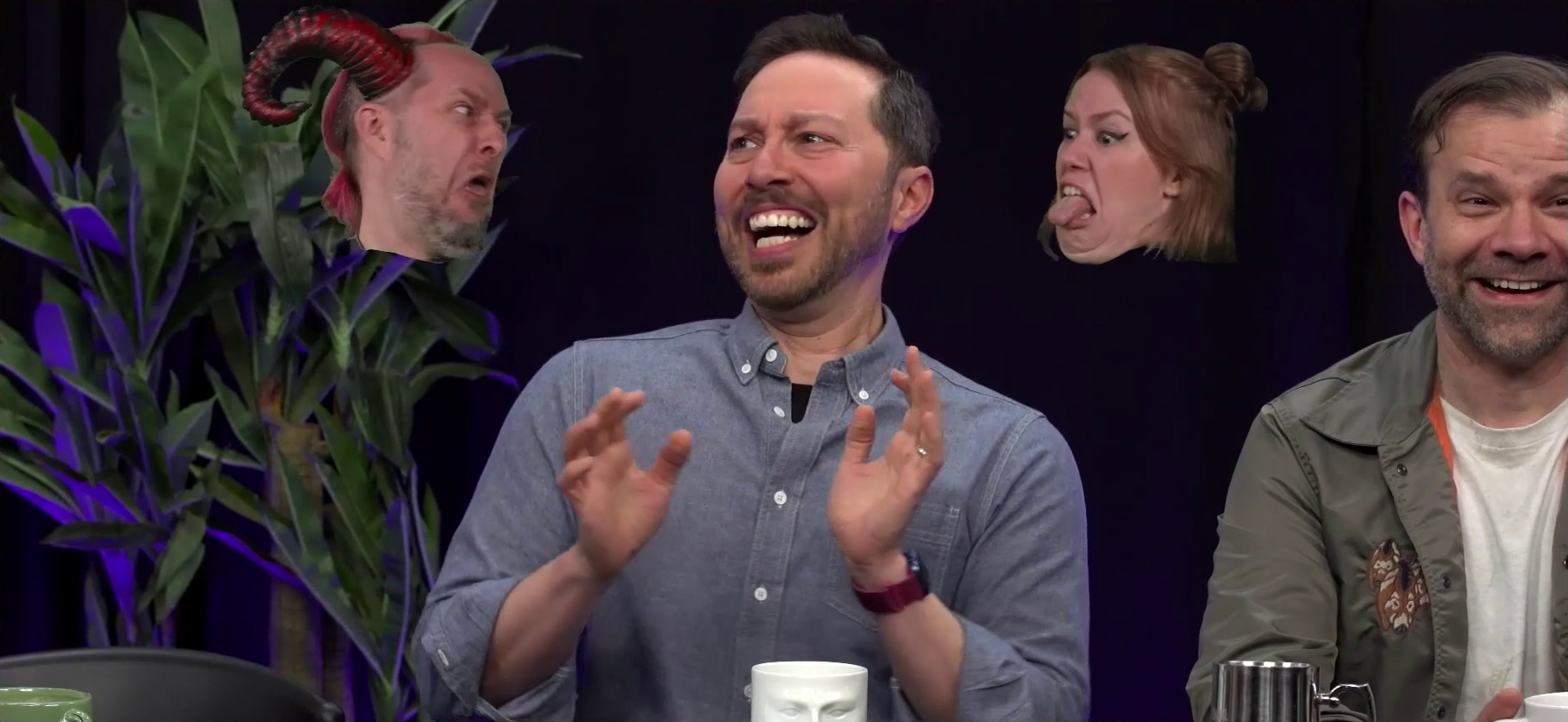 Sam at the Critical Role Announcement Playhouse table, with the floating head of Taliesin dressed like a devil over his right shoulder, and the floating head of Marisha with her hair in a bun over his left shoulder. Taliesin and Marisha are making frightening faces, Taliesin yelling and Marisha sticking out her toungue fiendishly. Sam looks frightened. Two red horns curl out of Liam’s head. Liam sits next to Sam, laughing.