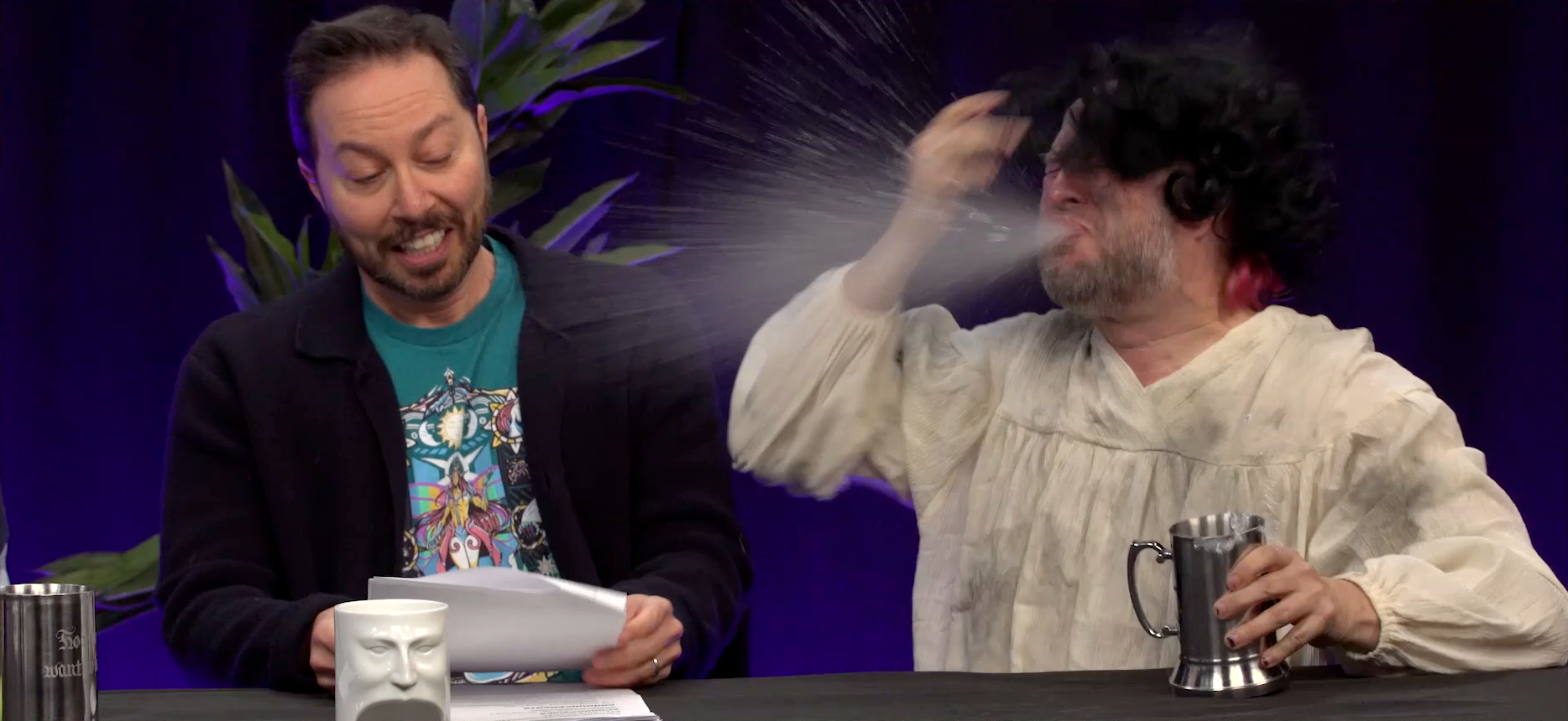 Sam and Taliesin at the Critical Role Announcement Playhouse table. Taliesin spits out liquid from his mouth in a wide spray directed at Sam. Taliesin is dressed like a medieval peasant, wearing a dirty white tunic and messy black wig over his real pink hair. Sam reads calmly from a stapled sheet of paper, not yet noticing the liquid headed his way. In Taliesin’s left hand is a metal stein.