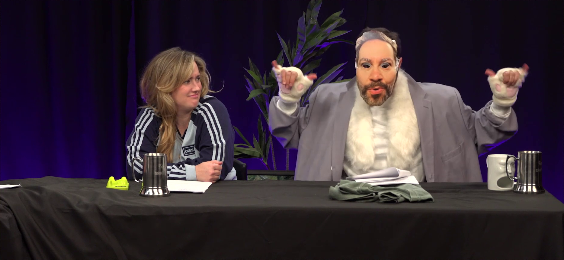 Ashley and Sam at the Critical Role Announcement Playhouse table. Sam wears a paper mask of his own face with eye holes cut out, a baggy gray blazer over a fuzzy white vest, and fuzzy white paw gloves. He flashes two hang-loose signs. Ashley looks over at him with confusion. Barely visible under the mask of Sam’s own face is a white mask.