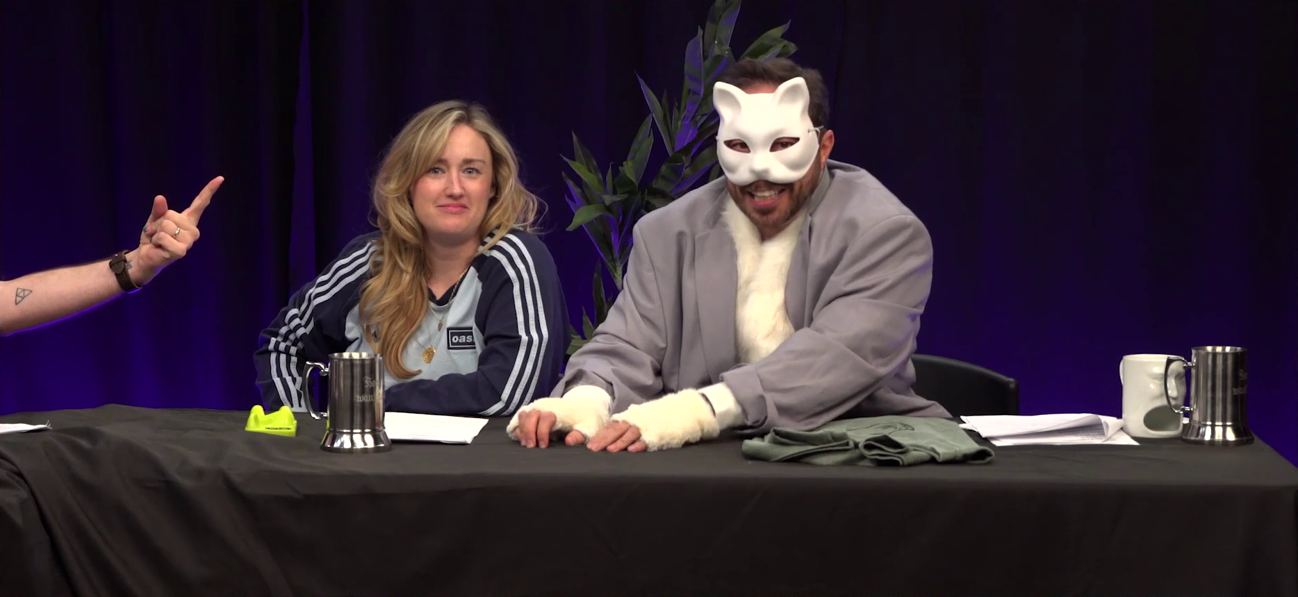 Ashley and Sam at the Critical Role Announcement Playhouse table. Sam wears a white cat mask, a baggy gray blazer over a fuzzy white vest, and fuzzy white paw gloves. He paws at the table in front of him, an apologetic look on his face. Ashley looks directly at the camera nonplussed. Matt’s hand comes in from out-of-frame, pointing one finger threateningly in Sam’s direction.