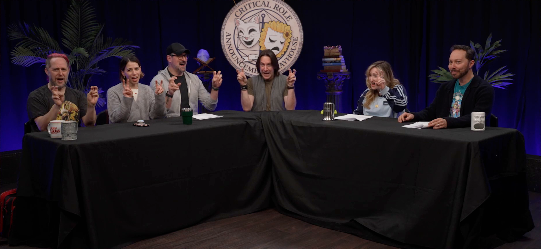 The Critical Role Announcement Playhouse table. Taliesin, Laura, Travis, Matt, and Ashley all make air quotes in unison. Sam does not, staring bemusedly at the camera with both his hands on the table in front of him.
