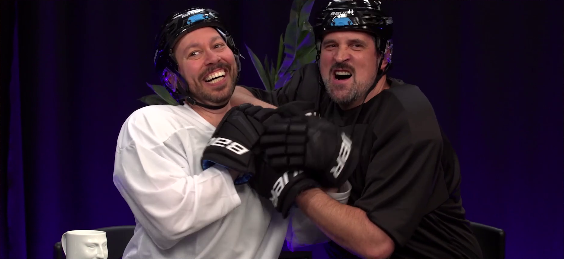 Sam and Travis at the Critical Role Announcement Playhouse table, dressed in hockey outfits and pawing at each other with large cumbersome hockey gloves. Sam is smiling goofily and laughing, while Travis makes a mock roaring face at the camera. Their gloves are tangled together in a way that indicates they are not sure what they’re doing.