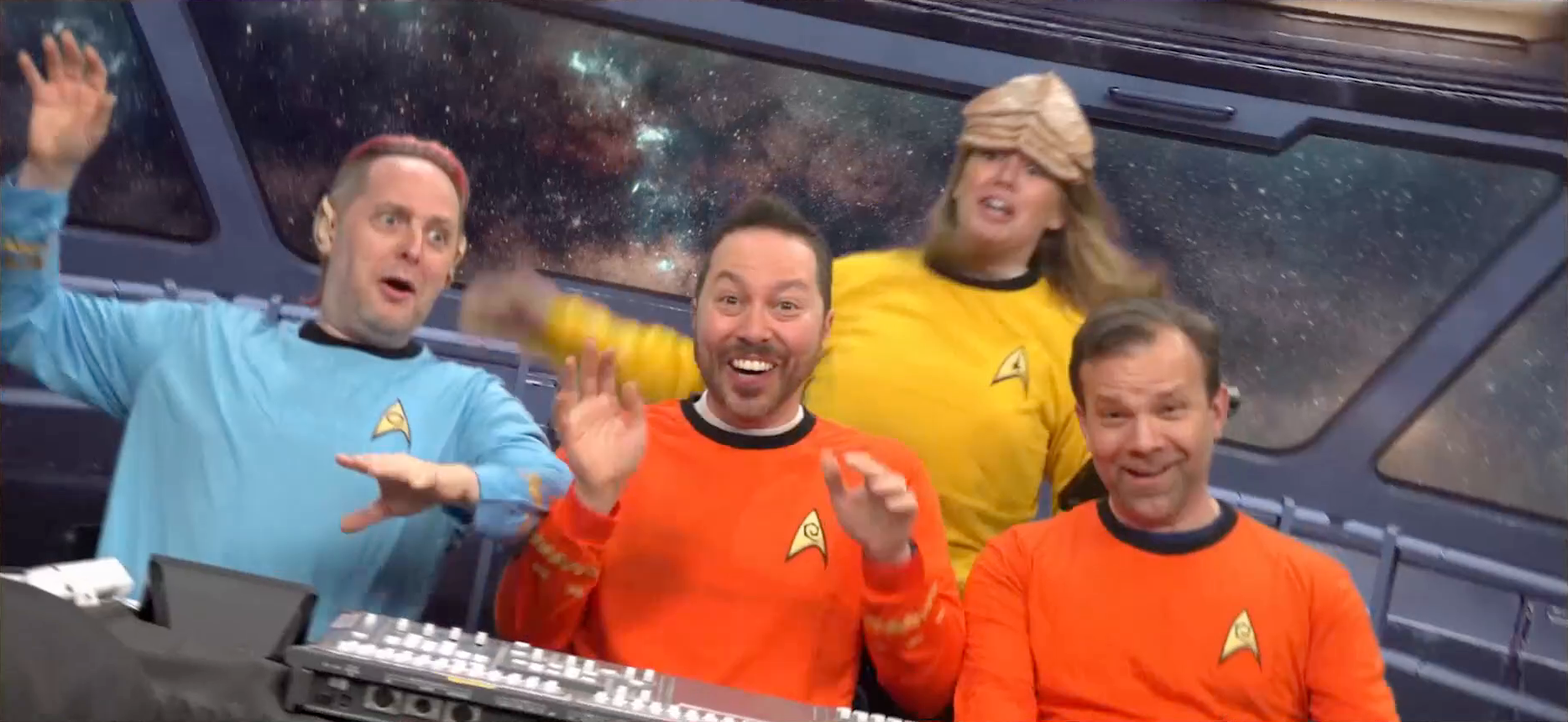 Taliesin, Sam, Marisha, and Liam all dressed in Star Fleet uniforms on a green-screened star ship bridge. The camera is tilted to indicate the ship is shaking, and the cast is pretending to be thrown around. Taliesin wears a blue shirt and Spock ears. Sam and Liam wear red shirts. Marisha wears a yellow shirt and a thick leathery alien forehead prosthetic. Taliesin and Marisha look scared, while Sam and Liam laugh. A sound mixing board resembling a control panel sits in front of Sam.