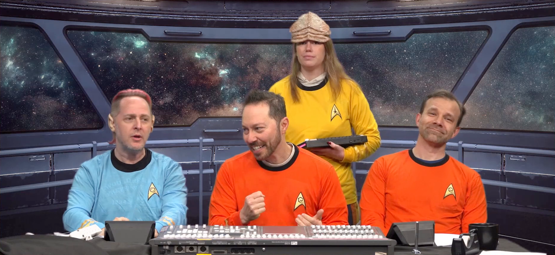 Taliesin, Sam, Marisha, and Liam all dressed in Star Fleet uniforms on a green-screened star ship bridge. Taliesin wears a blue shirt and Spock ears. Sam and Liam wear red shirts. Marisha wears a yellow shirt and a thick leathery alien forehead prosthetic. Sam clenches his fist dramatically in a Kirk-like manner, while Taliesin explains something, Liam mugs directly at the camera, and Marisha looks at the camera blankly. A sound mixing board resembling a control panel sits in front of Sam, along with a power strip, tape, a mug, and a couple small monitors.