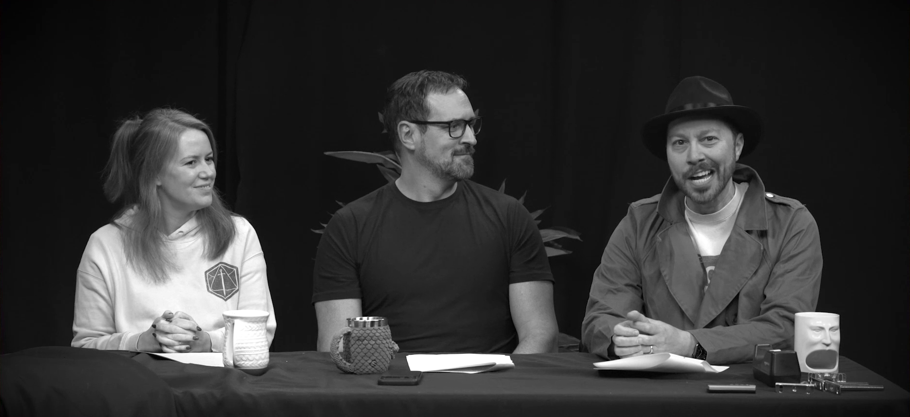 A black-and-white image of Marisha, Travis, and Sam at the Critical Role Announcement Playhouse table. Sam is dressed like a film noir detective, wearing a fedora and a trench coat over a white T-shirt with a partially visible graphic on the front. Sam speaks out of the side of his mouth, clearly doing a tough guy accent. Travis looks bemused. Marisha smiles and looks to the side, slightly confused, her hands clasped in front of her.