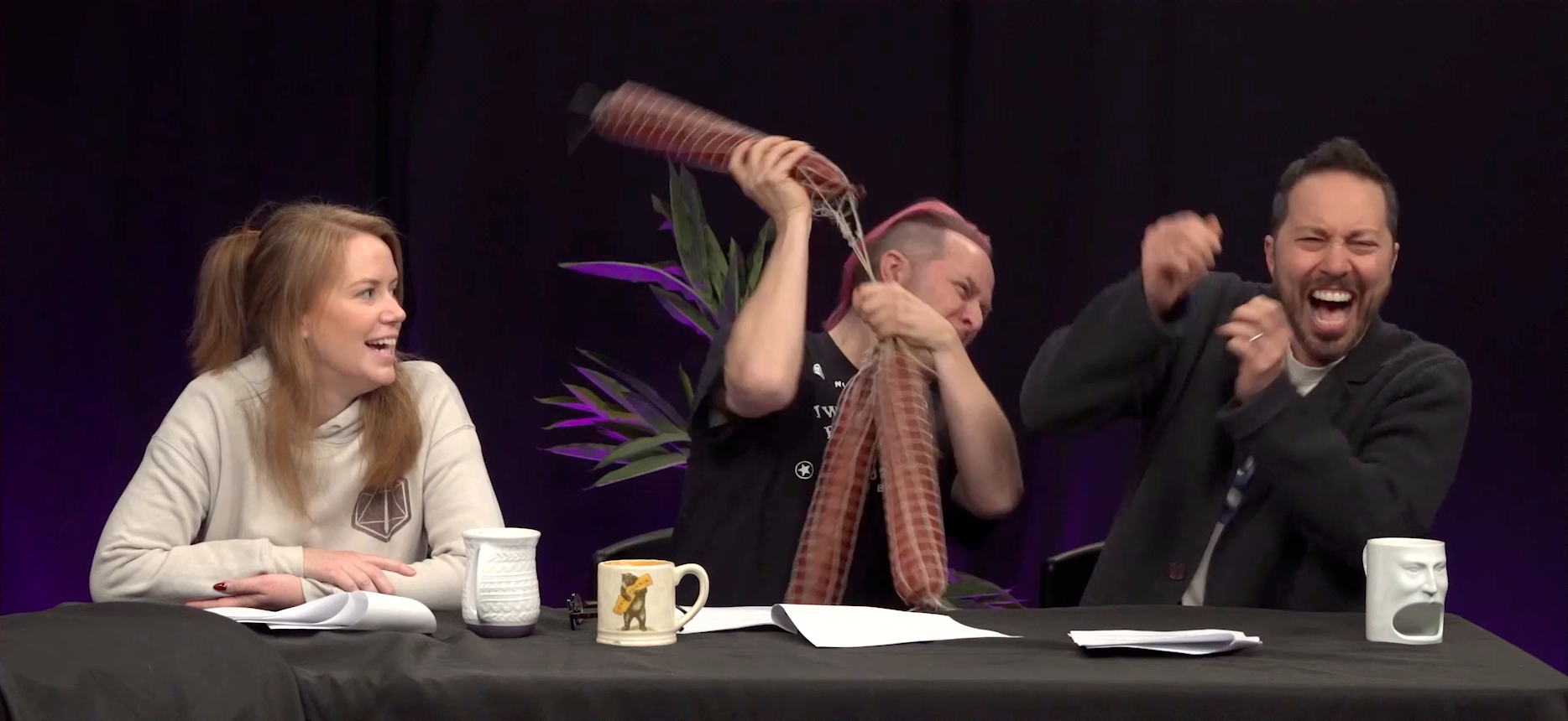 Marisha, Taliesin, and Sam at the Critical Role Announcement Playhouse table. Taliesin squints with effort as he swings back a large salami, ready to hit Sam. Sam raises his hands to protect himself and screams. Two other salamis, wrapped in netting, hang below the one being swung. Marisha looks on grinning, slight worry tinging the glee in her eyes.