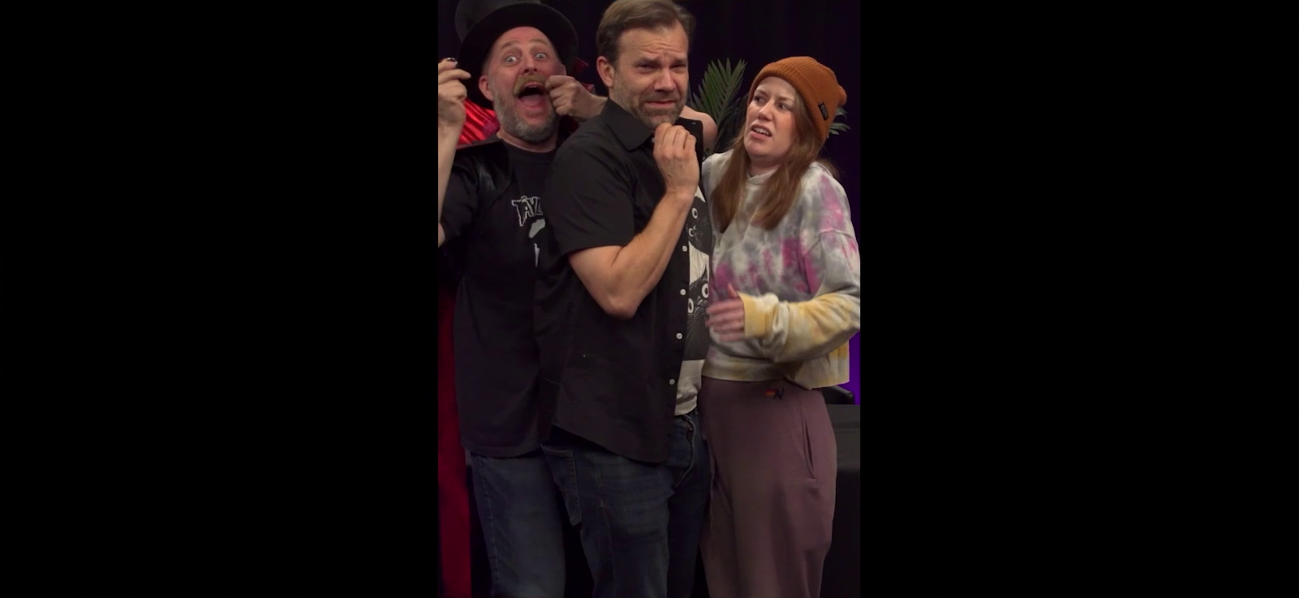 Taliesin, Liam, and Marisha squished into a vertical phone video frame, with large black bars on the left and right. Taliesin laugh maniacally, wearing a top hat and red cape as an old-timey villain costume. He holds a fake mustache over his face with his left hand and a wedding ring in his right hand. Liam and Marisha stand uncomfortably close to him and each other, struggling to fit in frame.