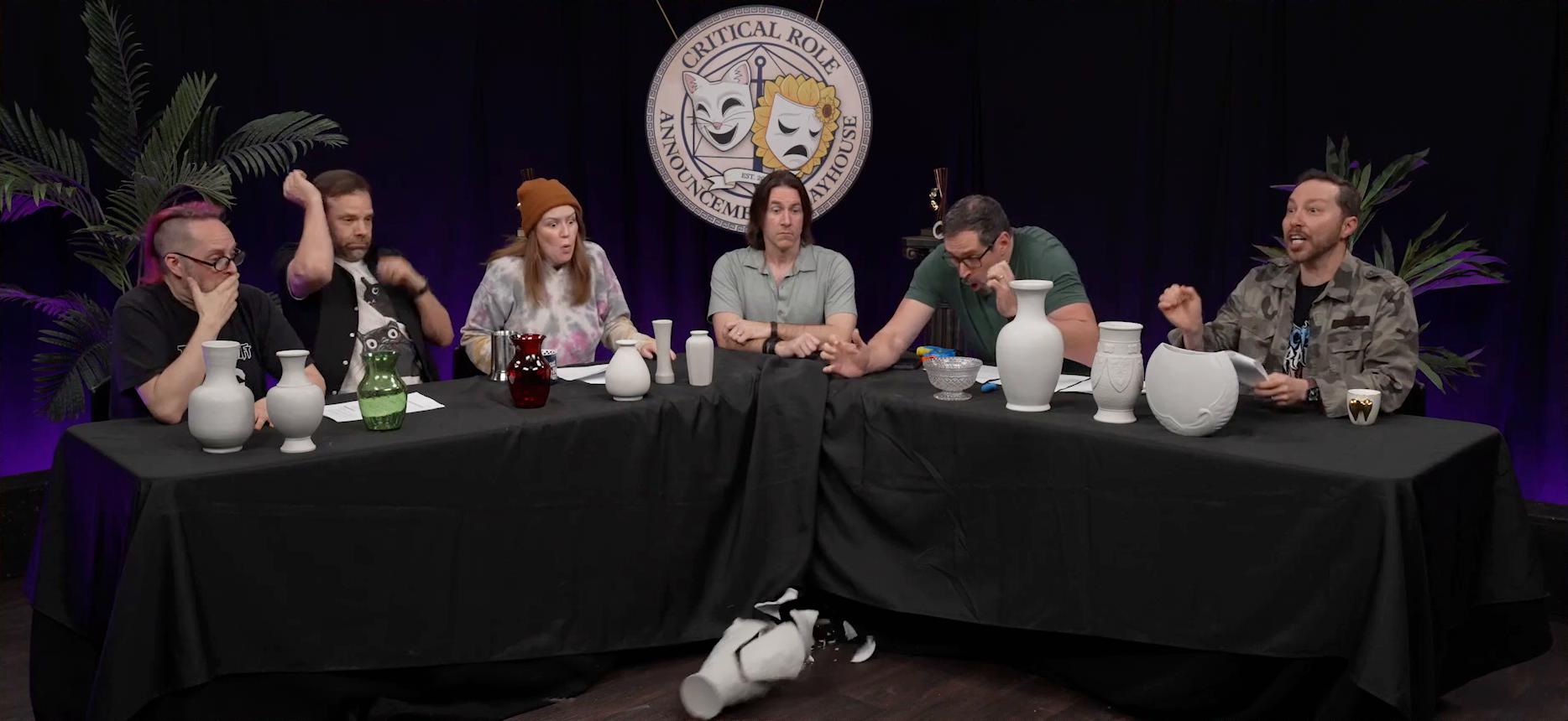 The Critical Role Announcement Playhouse table. Travis clumsily knocks a blank white vase off of the table. His face is a mask of surprise and regret as it smashes on the ground. Eleven other pieces of pottery, most white, one translucent green, and one translucent red, are arrayed across the table. Taliesin, Liam, Marisha, Matt, and Sam look on. Most look surprised, while Sam stops in the middle of reading from a script.