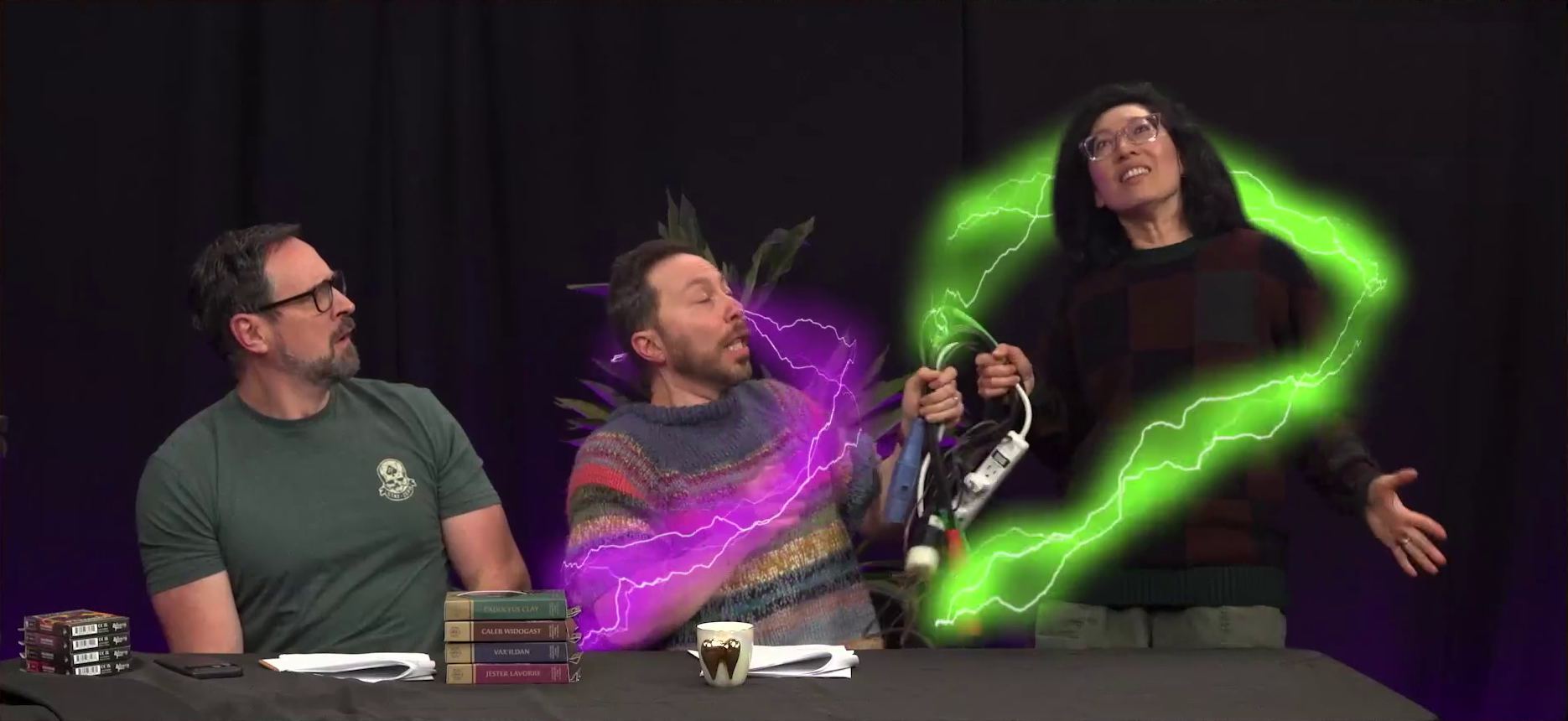 The Critical Role Announcement Playhouse table. Sam and Quyen have green and purple lightning surrounding them as they both hold on to an overloaded powerstrip. Their faces show a mix of pain and ecstacy. Travis looks on in confusion. On the table in front of them are four cardboard dice boxes labeled “VAX’ILDAN”, “CADUCEUS CLAY”, “JESTER LAVORRE”, and “CALEB WIDOGAST”.