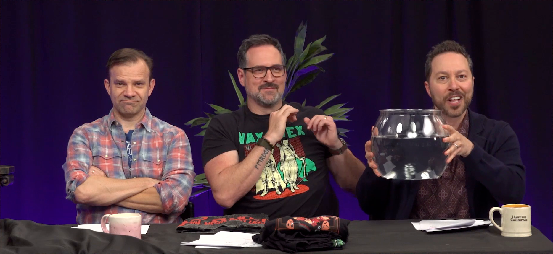 Liam, Travis, and Sam at the Critical Role Announcement Playhouse table. Sam holds up a round fish bowl filled with water and speaks. Travis leans away from him nervously. Liam looks directly into the camera, his face scrunched up in mock displeasure. Travis wears a shirt reading “VAX & VEX” with a monochrome image of Vax’ildan and Vex’ahlia on it.