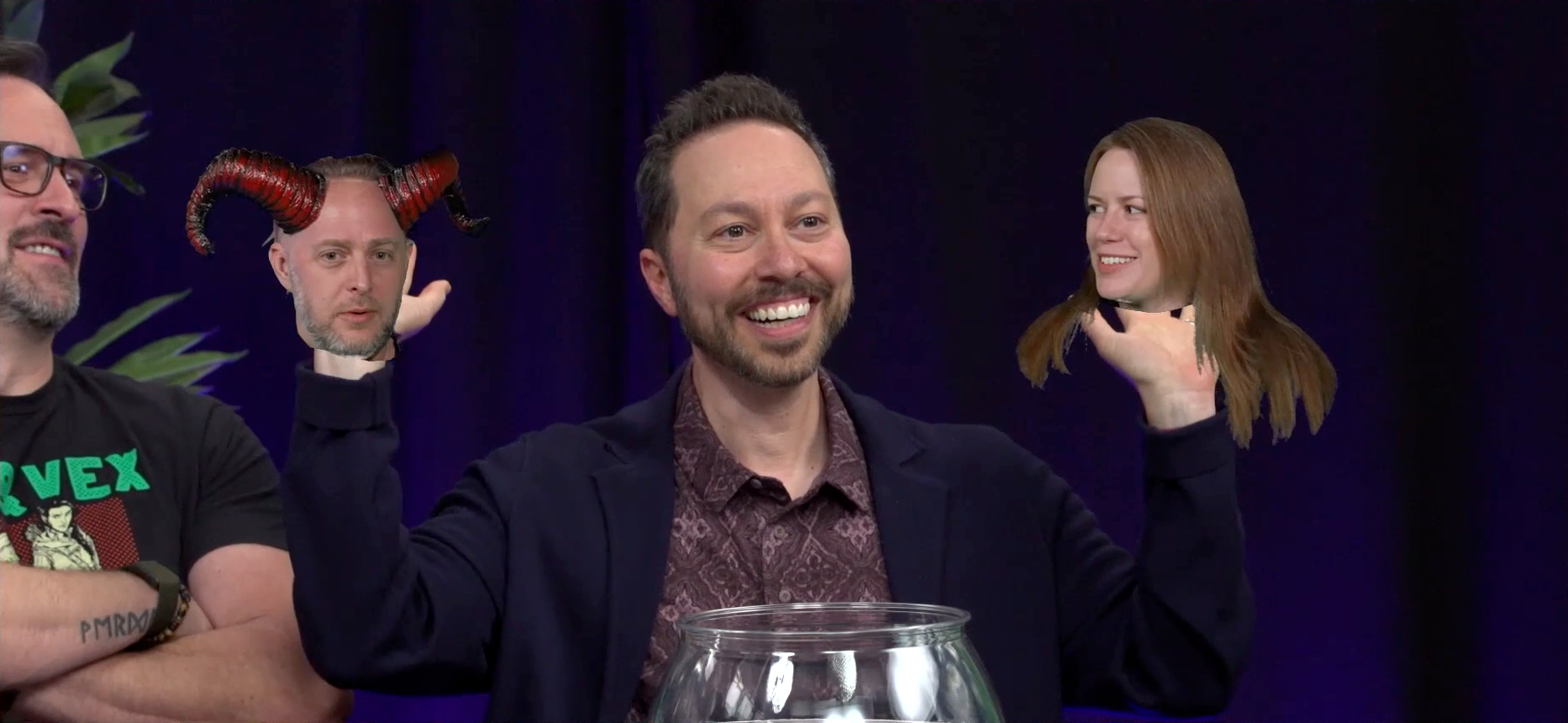 Sam at the Critical Role Announcement Playhouse table, with the floating head of Taliesin dressed like a devil over his right shoulder, and the floating head of Marisha over his left shoulder. Sam raises his hands behind the heads as if holding them up. Two red horns curl out of Taliesin’s head. He looks directly into the camera, vaguely bored. Marisha smiles and looks at Sam. Her hair trails below her floating head, resting on invisible shoulders. In front of Sam on the table is a round fish bowl filled with water. Travis sits next to Sam, laughing.