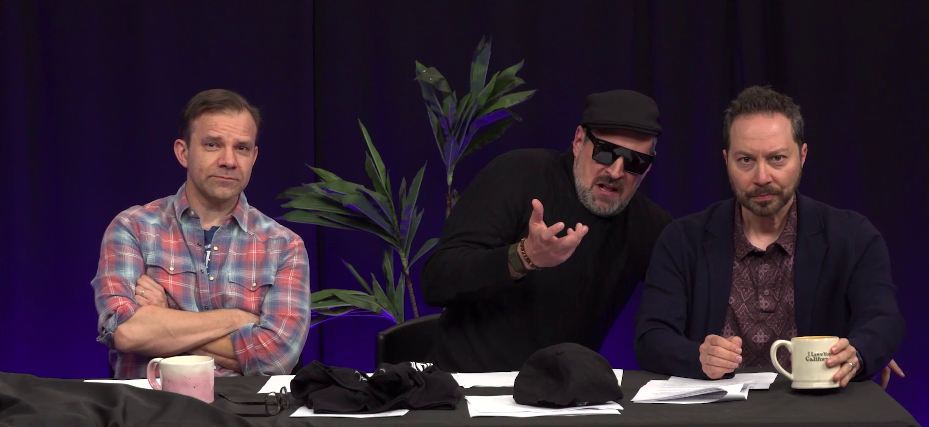 Liam, Travis, and Sam at the Critical Role Announcement Playhouse table. Travis is dressed like an artsy director in a black turtleneck, black beret, and sunglasses. He gestures in front of him, explaining something to Sam. Sam looks attentive. Liam seems slightly amused, looking directly into the camera.