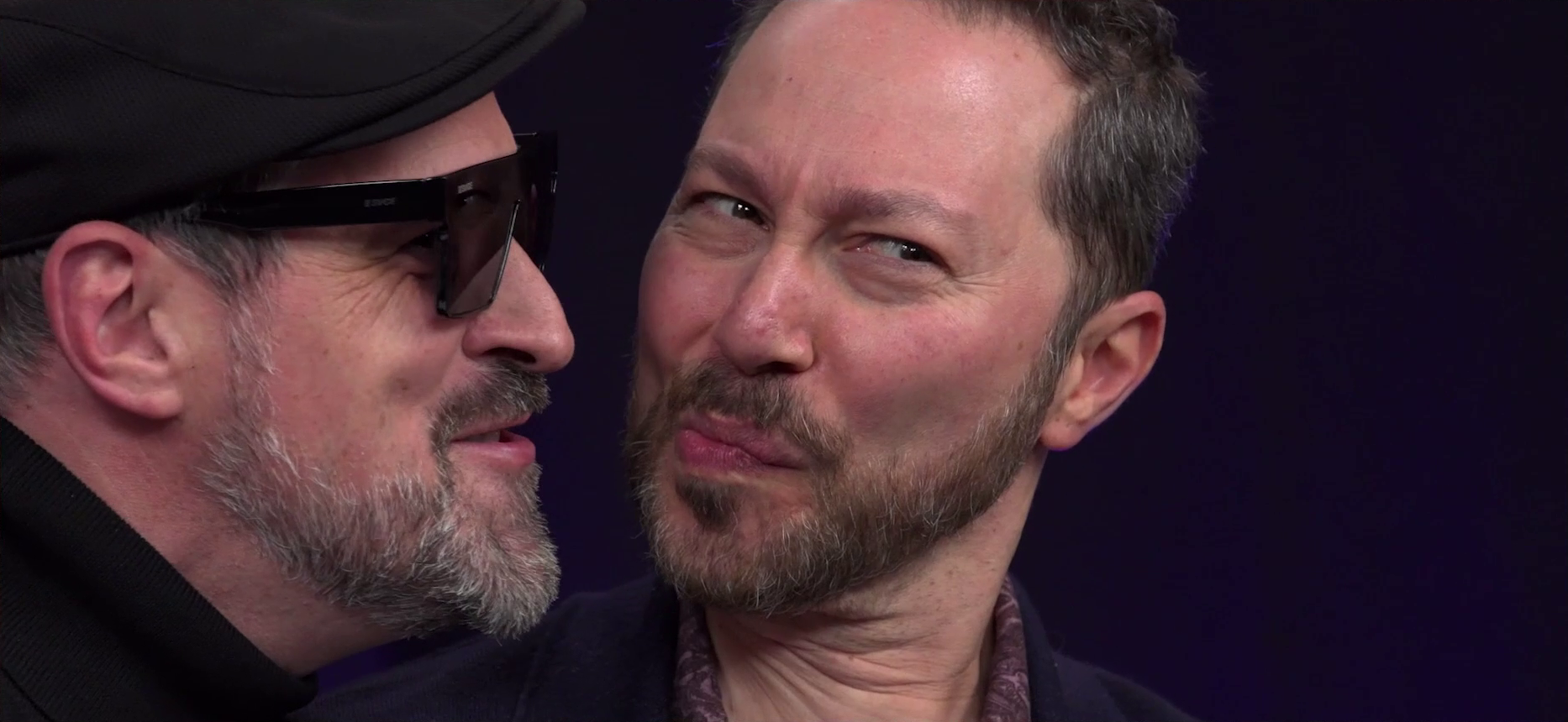 Travis speaking with his face inches away from Sam’s. Sam purses his lips to the side into a sneaky kiss, while Travis struggles not to smile. Travis is dressed like an artsy director in a black turtleneck, black beret, and sunglasses.
