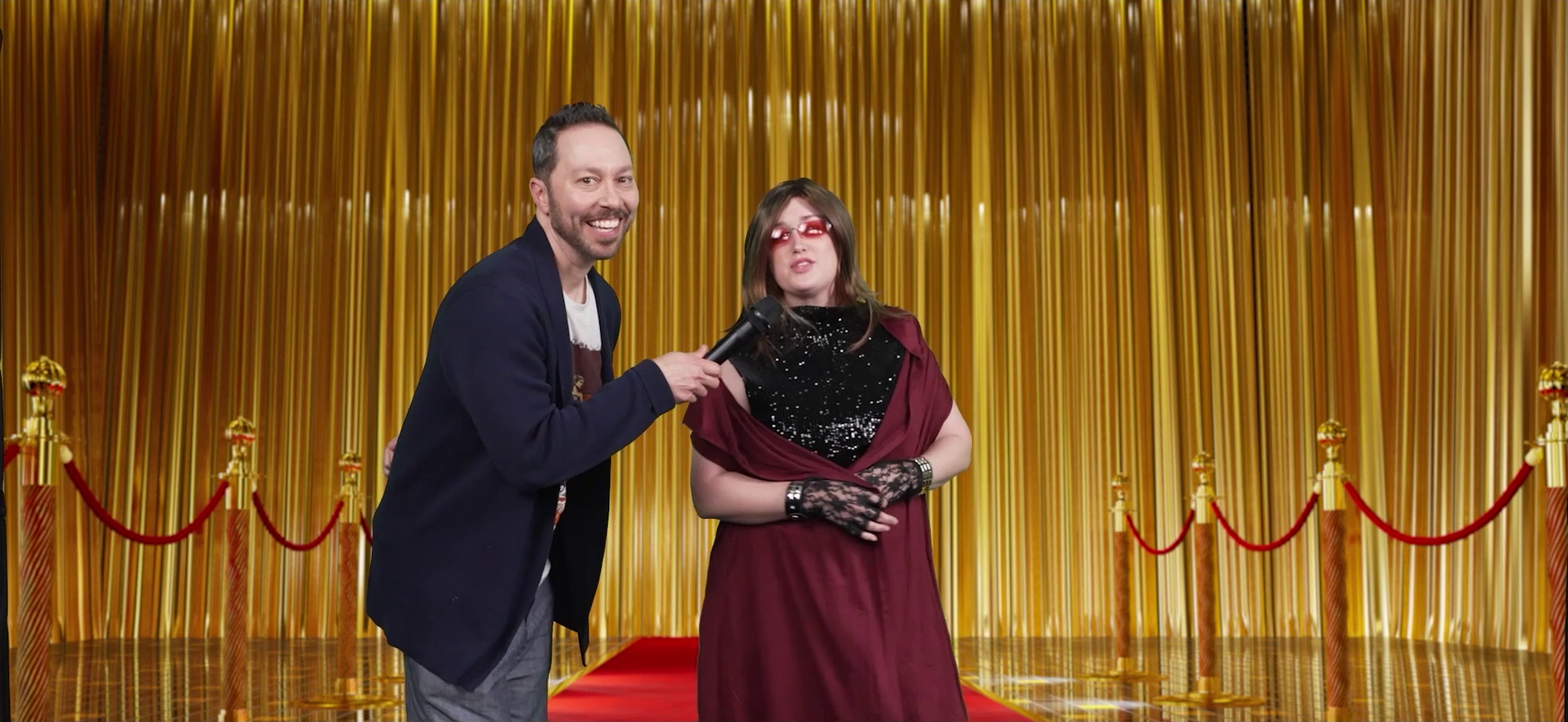 Sam wearing a blazer interviewing a fancily-dressed Ashley on a red carpet. Ashley has a brown wig just past her shoulders, red-lensed glasses, a sparkly black gown, and a sleeveless vest-like red shawl pulled over it. She wears lacy black gloves and studded punk bracelets. Ashley talks into Sam’s microphone while Sam smiles at the camera. A golden curtain is in the background.