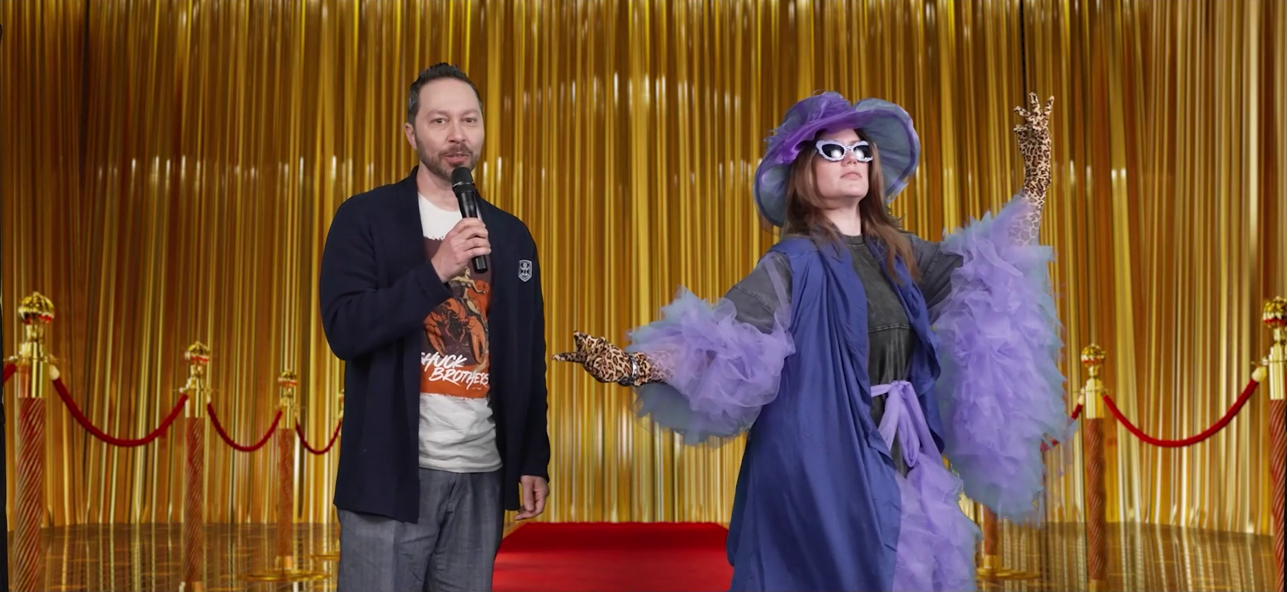 Sam wearing a blazer interviewing a fancily-dressed Marisha on a red carpet. Marisha has a purple wide-brimmed hat covered in toile, silver-rimmed sunglasses, a long purple vest-like jacket, poofy purple toile sleeves, and leopard print gloves. She strikes a dramatic pose with her arms out while Sam speaks into a microphone, looking deadpan at the camera. A golden curtain is in the background.