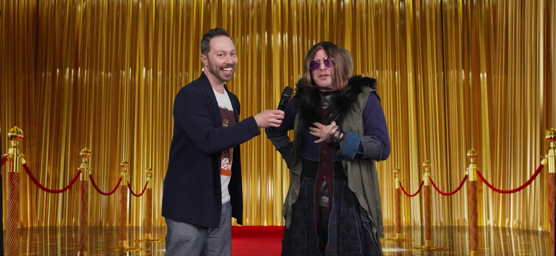 Sam wearing a blazer interviewing a fancily-dressed Taliesin on a red carpet. Taliesin has a brown wig just past his shoulders, round purple-lensed glasses, a sleeveless vest-like brown jacket with a black fur collar, a roughly-knit purple sweater that goes down past his knees, and a number of thin bracelets. He speaks into Sam’s microphone, looking serious while Sam smiles at the camera. A golden curtain is in the background.