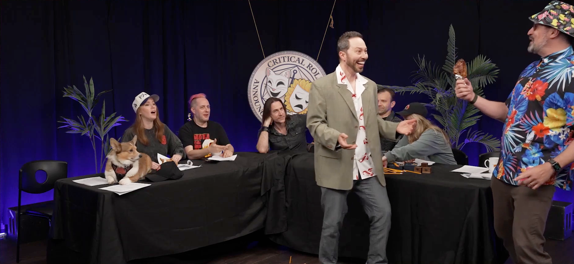 The Critical Role Announcement Playhouse table. Sam and Taliesin stand in front of it acting out a scene. Sam is wearing a tan blazer over a white button-up with images of lobsters on it. Travis is wearing a Hawaiian shirt, floral bucket hat, and sunglasses, and is holding a turkey leg. Sam is smiling and looking surprised while Travis laughs genially. Omar the dog, Marisha, Taliesin, and Matt look on from the table bemused. Liam and Ashley are distracted cleaning up a bunch of spilled pencils on the table.