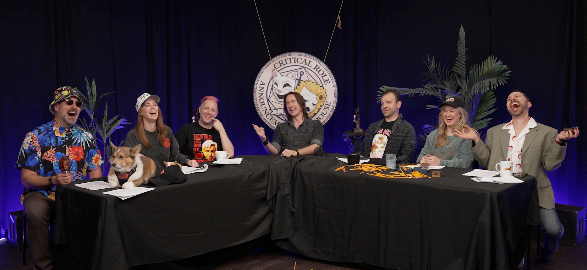 The Critical Role Announcement Playhouse table. Travis, Omar the dog, Marisha, Taliesin, Matt, Liam, Ashley, and Sam sit laughing, as if posed. Travis is wearing a Hawaiian shirt, floral bucket hat, and sunglasses, and is holding a turkey leg. Sam is wearing a tan blazer over a white button-up with images of lobsters on it and is holding a wooden ring box. A bunch of pencils are spilled on the table in front of Liam, Ashley, and Sam.