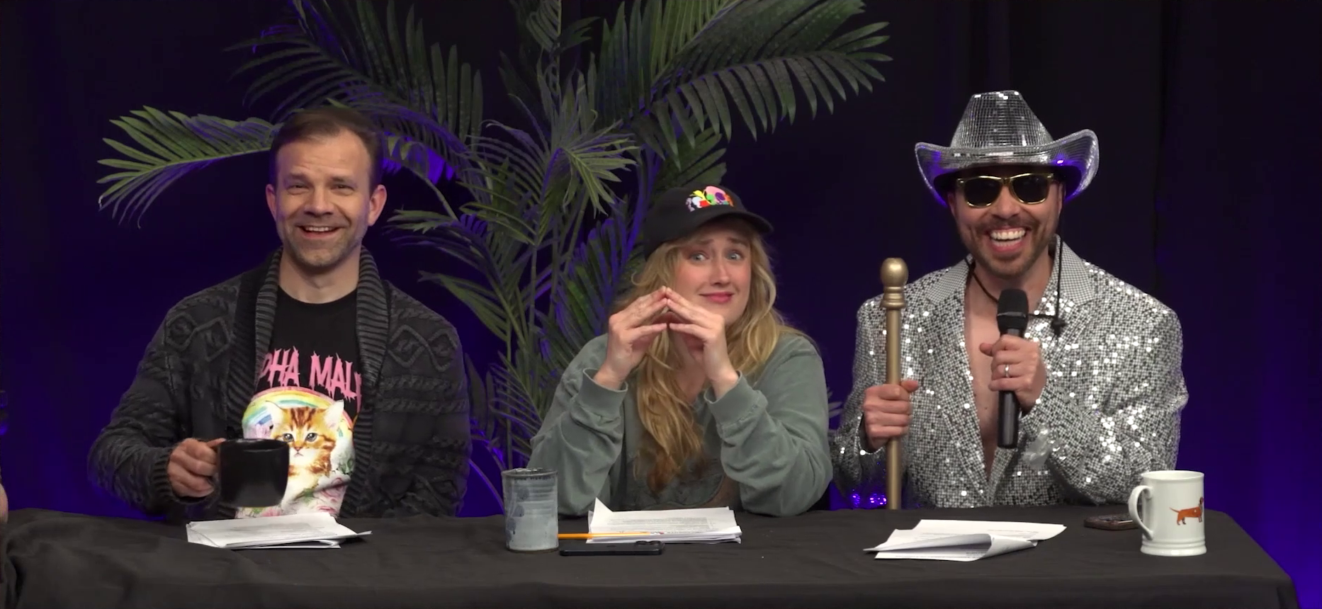 Liam, Ashley, and Sam sit at the Critical Role Announcement Playhouse table. Sam is dressed in a silver sparkly sequined blazer and cowboy hat, similar in material to the surface of a disco ball. He holds a microphone in his left hand and a long golden staff in his right hand, and smiles broadly under his sunglasses. He is not wearing a shirt under the blazer and it is open below his chest. Liam grins affably, holding a mug of coffee in his hand. Ashley steeples her fingers, looking directly at the camera with a goofily frightened expression on her face.