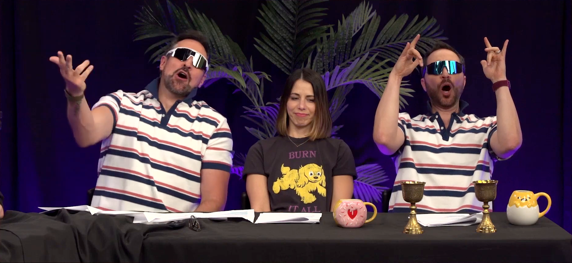 Travis, Laura, and Sam at the Critical Role Announcement Playhouse table. Travis and Sam are dressed as frat bros with striped red, white, and blue polo shirts and wrap-around sunglasses. They are both loudly shouting, Travis gesturing forward and Sam making “rock on” shapes with his hands. Laura sits between them, looking bored and uncomfortable. She is wearing a T-shirt with art of a yellow shaggy dog on the front and the text “BURN IT ALL”.