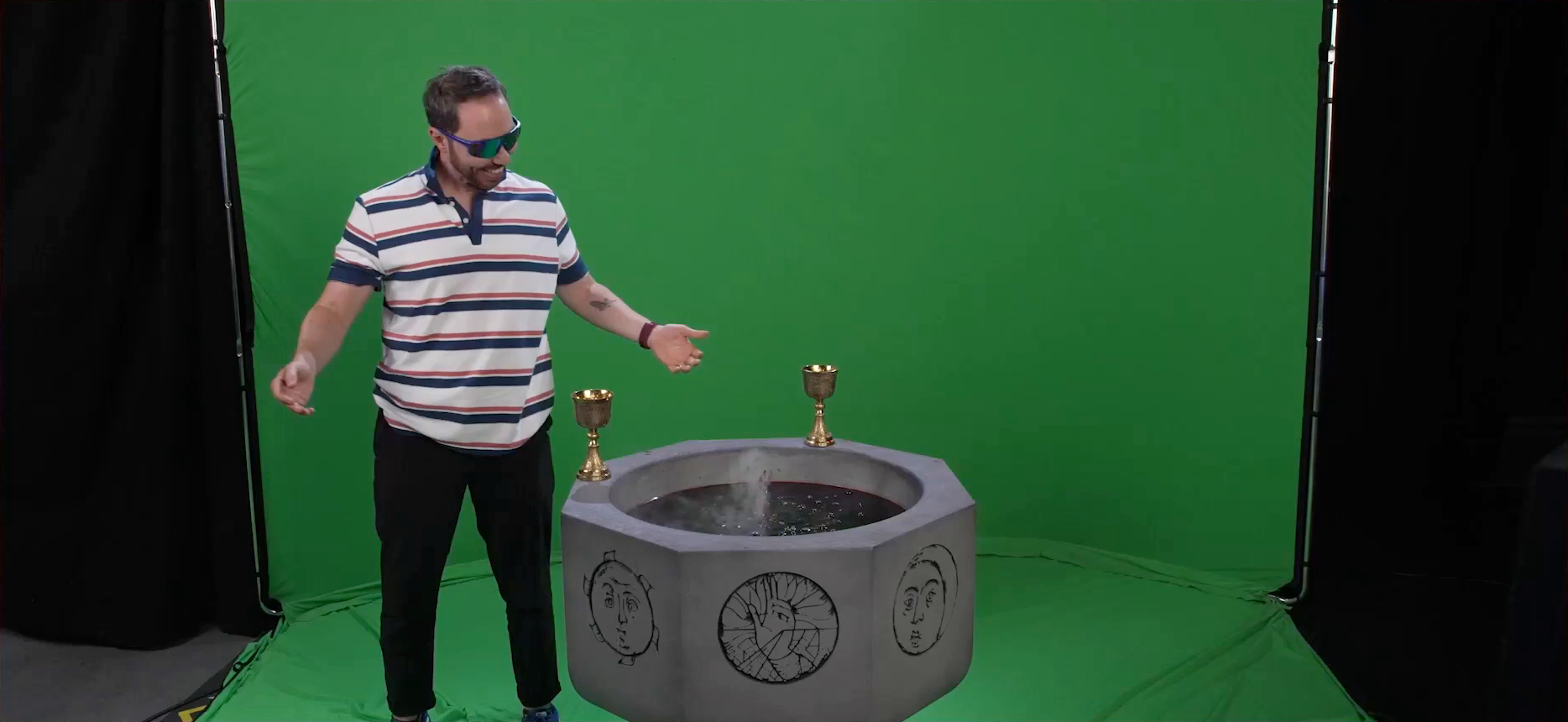 Sam standing next to a bubbling fountain of blood in front of a green screen. He is dressed as a frat bro with a striped red, white, and blue polo shirt and wrap-around sunglasses. Two gold chalices sit on the rim of the stone fountain. Designs of a sun, a moon, and a hand in a benediction gesture are on the sides of the fountain. Sam smiles at the fountain and shrugs.