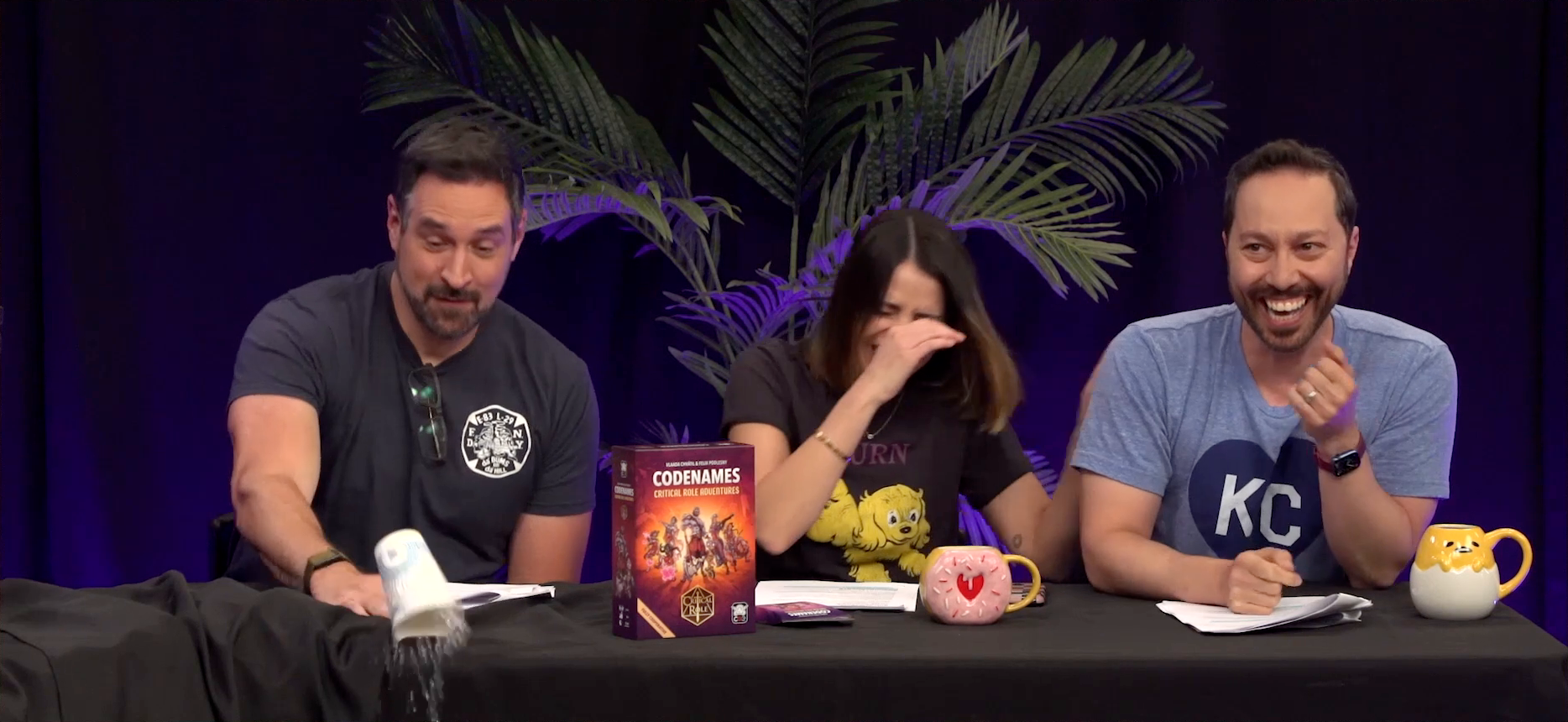 Travis, Laura, and Sam at the Critical Role Announcement Playhouse table. Travis knocks a paper cup of water off the table while Laura and Sam laugh. Travis’s eyes are half closed as if having just woken from a doze. Laura half covers her face with her hand in laughter. Sam smiles widely, Laura’s hand on his back. A board game box labeled “CODENAMES: CRITICAL ROLE ADVENTURES” stands on the table in front of them.