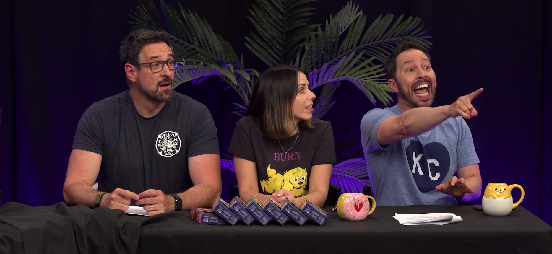 Travis, Laura, and Sam at the Critical Role Announcement Playhouse table. Sam points excitedly to something out of frame, his mouth open in a mixture of fear and surprise. Travis squints his eyes looking where Sam is pointing, his mouth slack. Laura looks at Sam with a mixture of interest and amusement. On the table are stacked seven small boxes of Vox Machina dice sets.