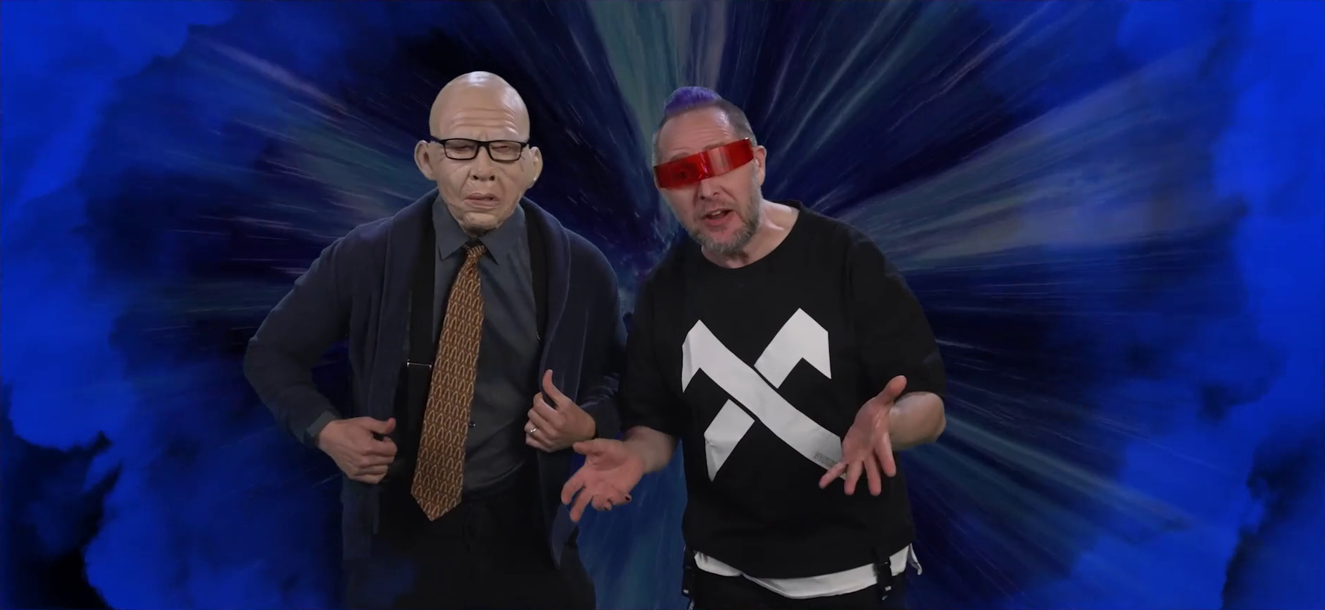 A future Sam and Taliesin standing in a time vortex made of blue light. Sam wears an old man mask that fully obscures his face, glasses, a dress shirt and tie, suspenders, and a dark blue cardigan. Taliesin wears a red-tinted futuristic visor over his eyes but otherwise doesn’t look any different than he normally does. Both are leaning forward looking into the camera, and Taliesin has his hands out as if explaining something.