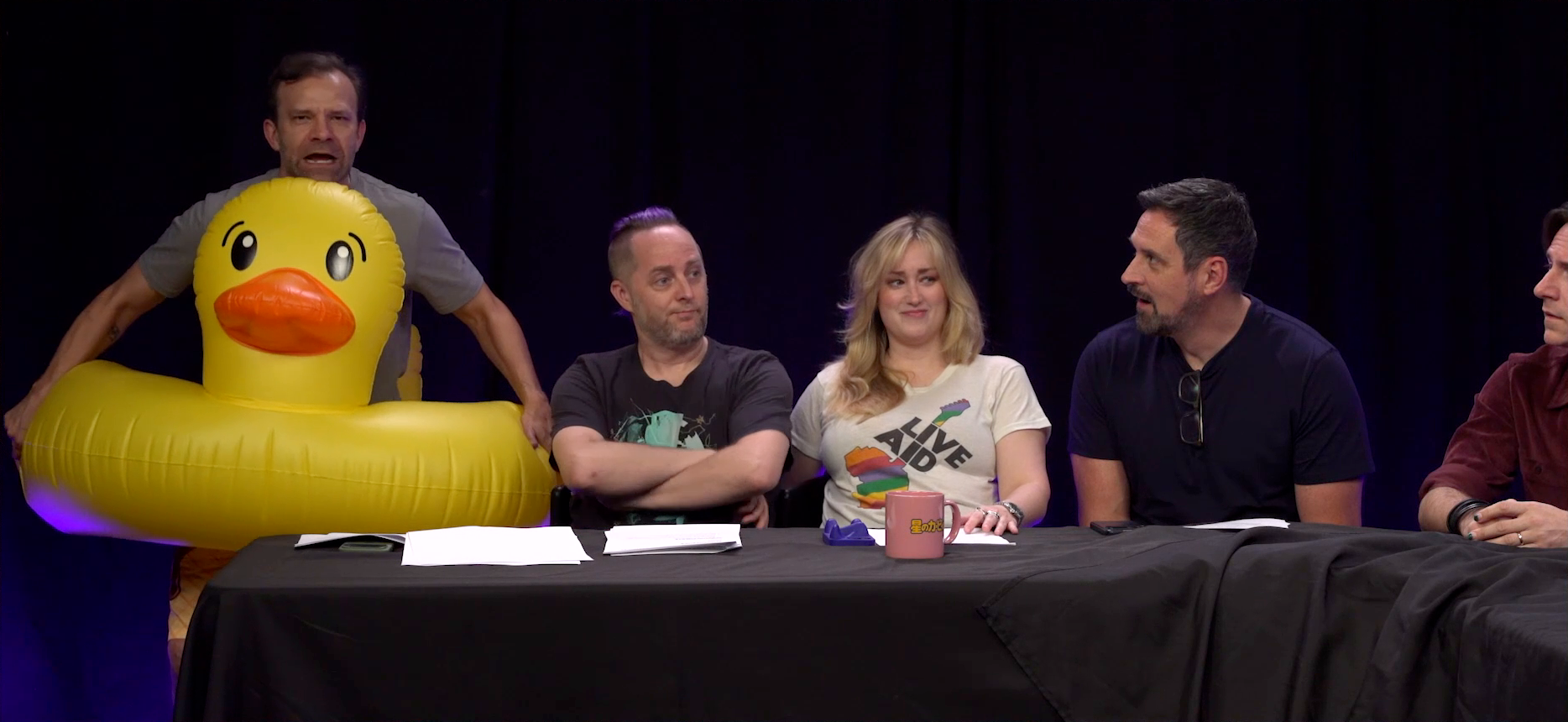 Liam, Taliesin, Ashley, Travis, and Matt at the Critical Role Announcement Playhouse table. Liam wears a giant inflatable duck floaty and yells directly into the camera. Taliesin looks away in disappointment, Ashley looks to her side amused, Travis looks at Liam mouth agape, and Matt looks at Liam confused.