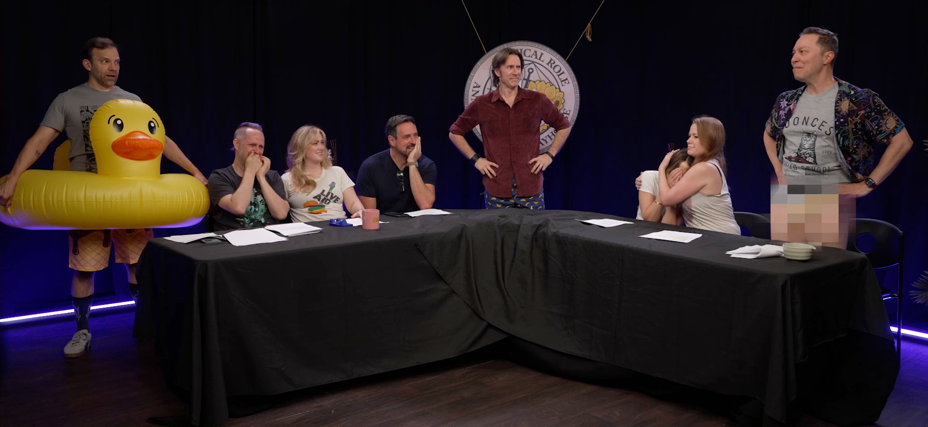 Sam at the Critical Role Announcement Playhouse table, standing up defiantly to reveal he’s not wearing any pants. A pixelated censor bar is over his crotch. Liam stands at the other end of the table in a giant inflatable duck floaty, looking at Sam in horror. Taliesin covers his mouth in laughter, his face red. Ashley smiles calmly at Sam. Travis laughs, looking at Sam with his chin resting on his palm. Matt, wearing navy blue boxers with bananas on them, looks at Sam perturbed. Laura covers her face in fear, and Marisha holds her to her chest as if protecting her.