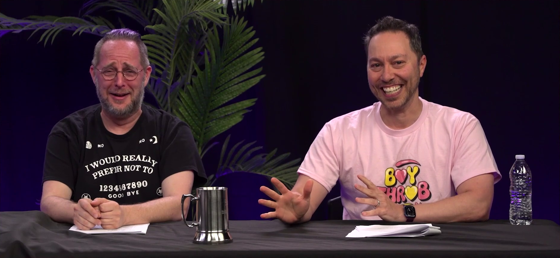 Taliesin and Sam at the Critical Role Announcement Playhouse table. Taliesin is red faced, crying laughing, as Sam smiles and tries to explain something to the camera. Sam’s hands are in front of him as if presenting a concept. Taliesin wears a shirt with an image of an Ouija Board on it, but with the option “I WOULD REALLY PREFER NOT TO” included. Sam wears a T-shirt with pink and yellow text reading “BOY THROB” with the letters “O” replaced with hearts.