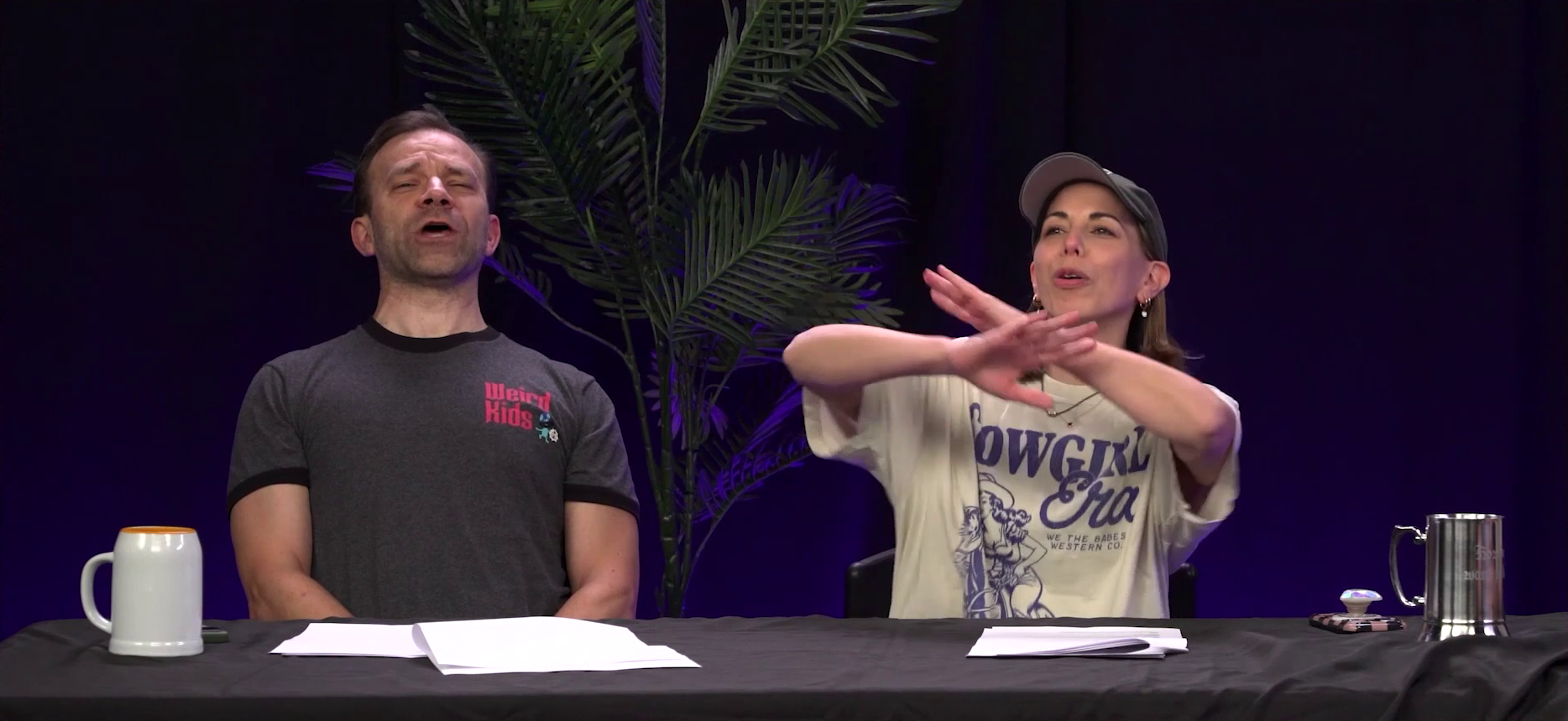 Liam and Laura at the Critical Role Announcement Playhouse table. Both are bombastically announcing something, like hype men. Liam leans back and shouts into the air. Laura crosses her arms in front of her emphatically while shouting. Liam wears a T-shirt with red left breast text reading “Weird Kids”. Laura wears a T-shirt with an image of a cowgirl on the front reading “COWGIRL Era. WE THE BABES WESTERN CO.”.