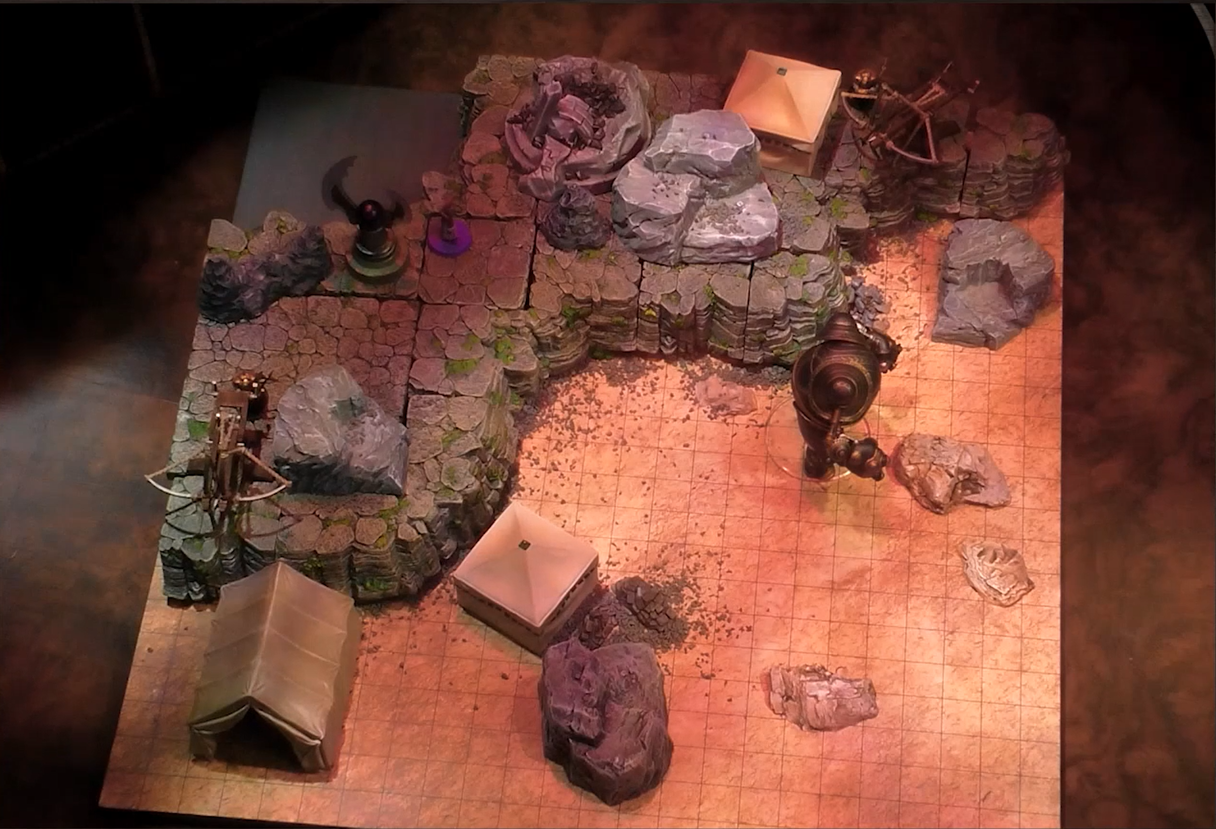 A battlemap with a raised natural rock surface in the corner. One square tent, two ballistas, a winged token, and Imogen sit on the surface and two other tents and a rounded robot sit on the lower ground.