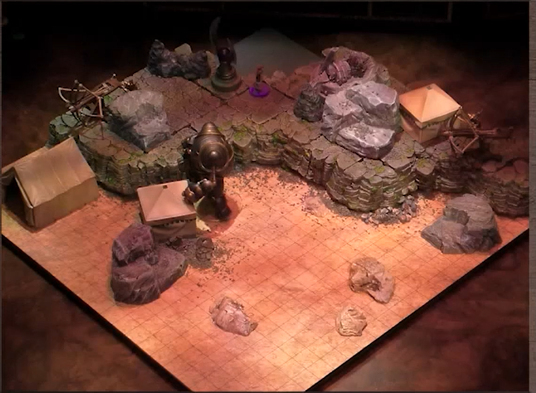 A battlemap with a raised natural rock surface in the corner. One square tent, two ballistas, a winged token, and a Imogen sit on the surface and two other tents and a rounded robot sit on the lower ground. The robot is standing over a tent.
