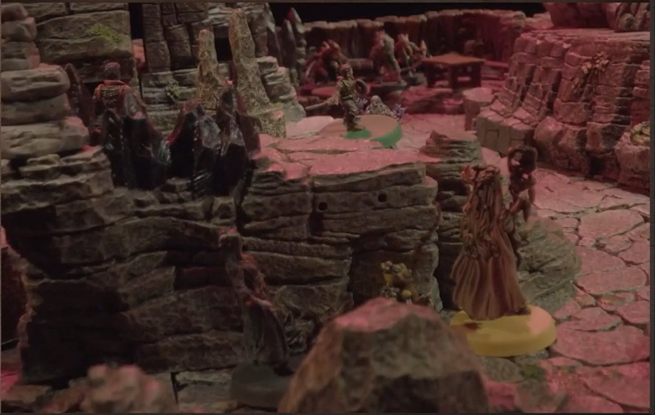 A battlemap of rock-walled space dotted with rock formations and stalagmites. Orym is on a ledge, Fearne is in the foreground near him, Imogen is visible in the background, and a humanoid with armor is on the ledge near Orym.