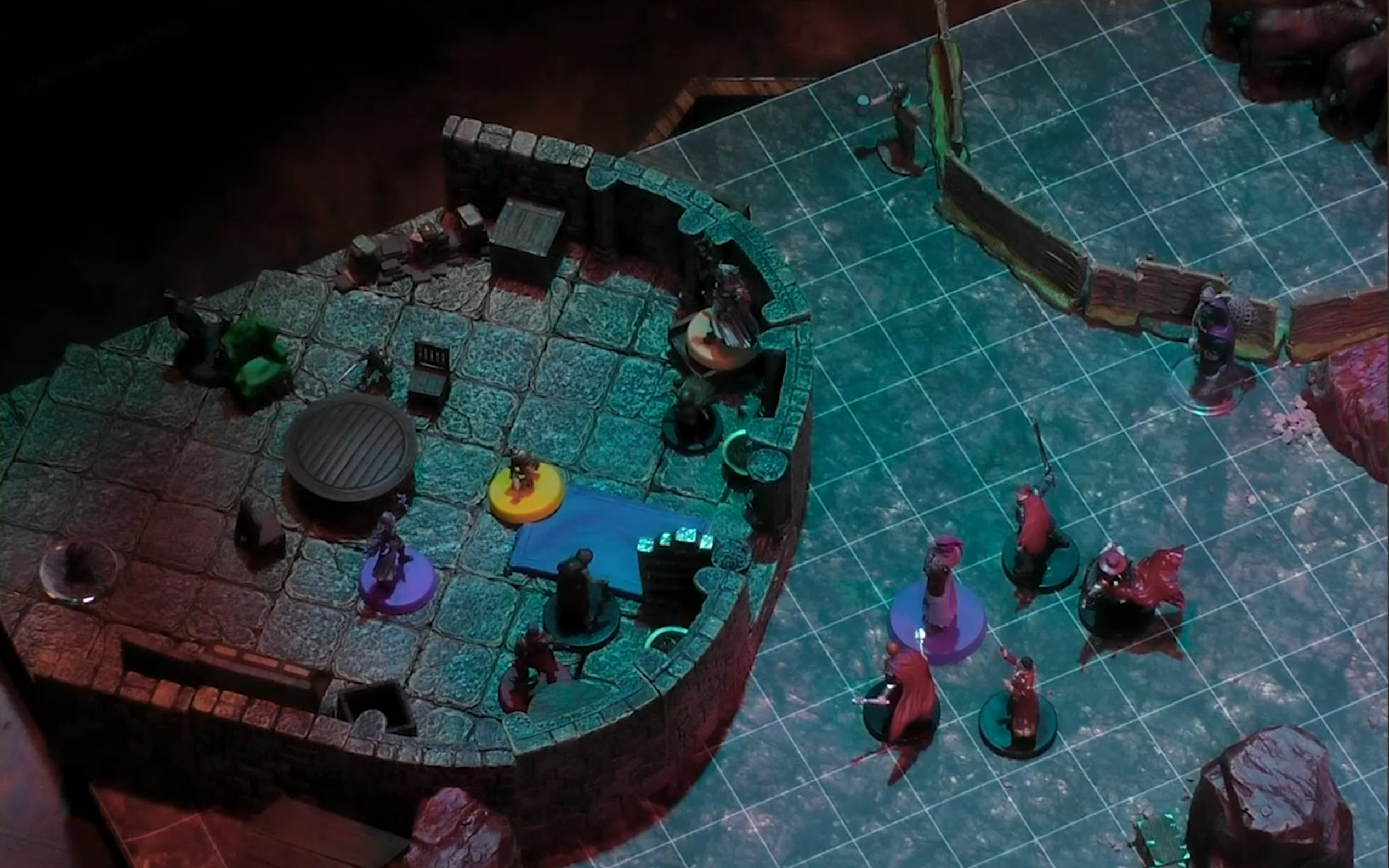 A battlemap of a rounded stone building with an expanse of flat ground outside. FCG, Orym, Imogen, Fearne (pressed against a wall), and other tokens are inside the building. A purple token stands outside at the entance surrounded by red-caped individuals. Two other tokens stand by a fenced area.