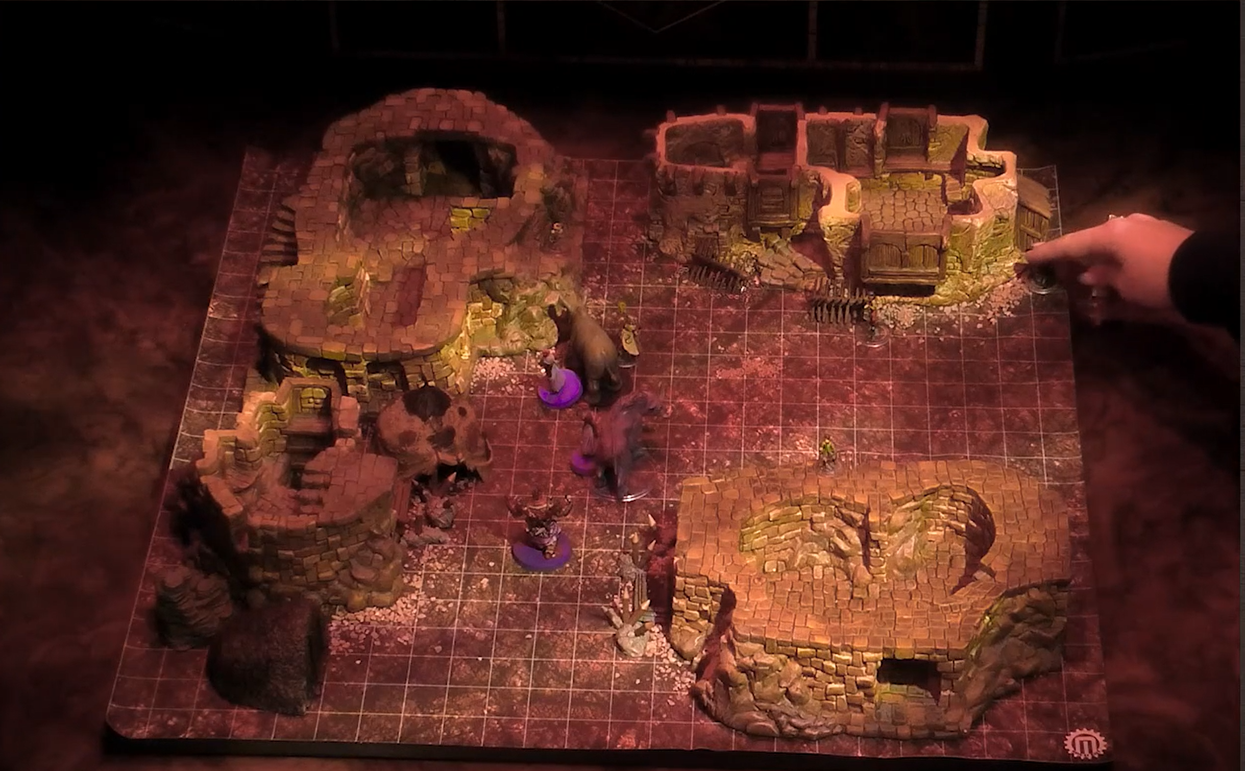 A battlemap centered on a street between four stone buildings. Four purple-based tokens with two large tusked quadripedal creatures are in the street. Orym is on the roof of a nearby building.